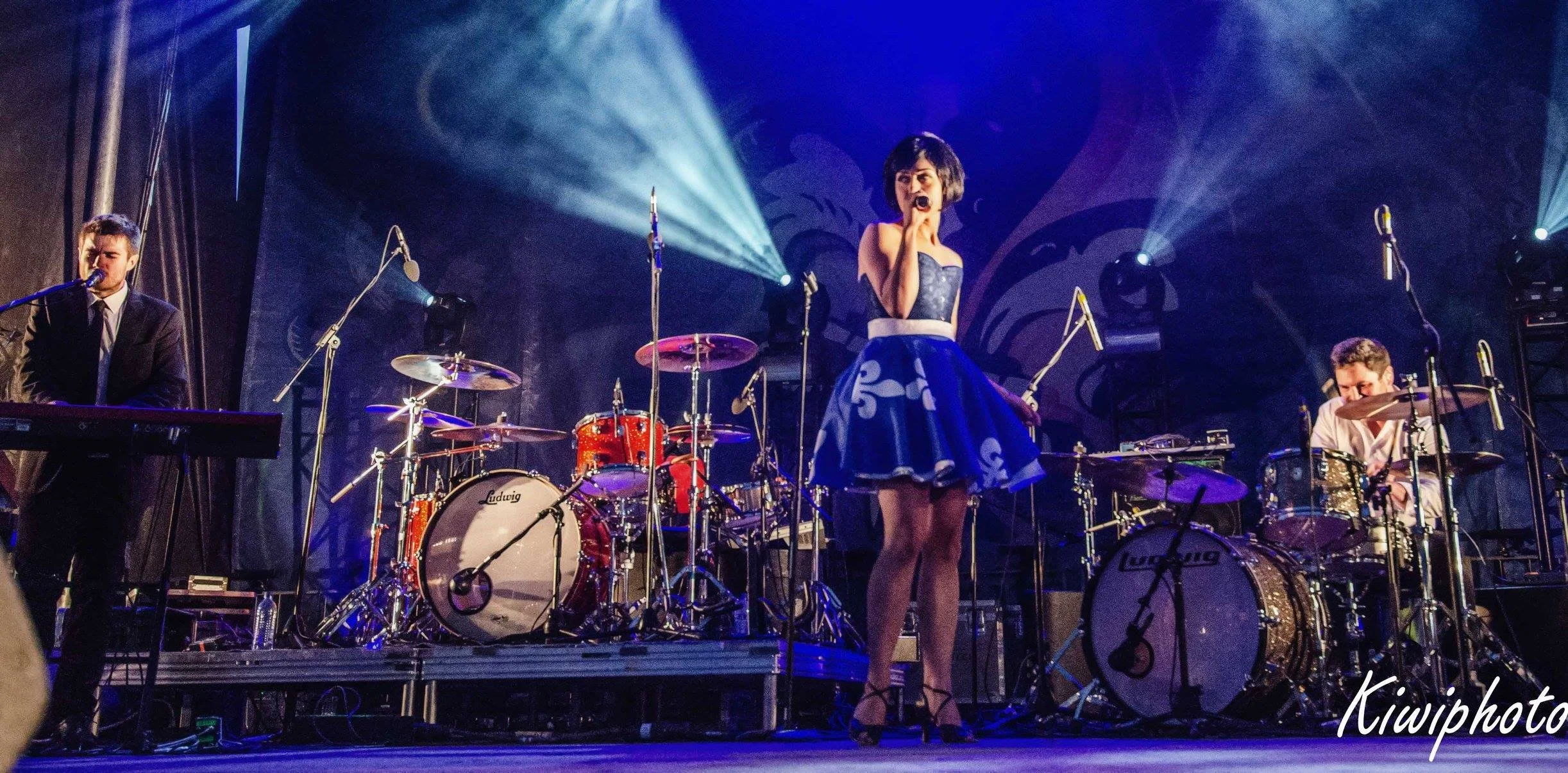 Une femme chante au centre d'une scène avec un groupe de musiciens derrière elle. La scène est éclairée par des projecteurs et la femme porte une robe bleue à motifs blancs.