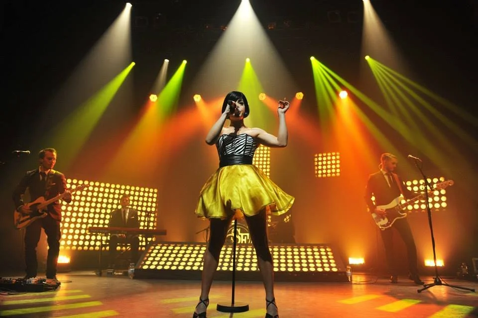 Une chanteuse en robe jaune et haut noir se produit sur scène avec un groupe musical, sous un éclairage coloré et des projecteurs en forme de carré.