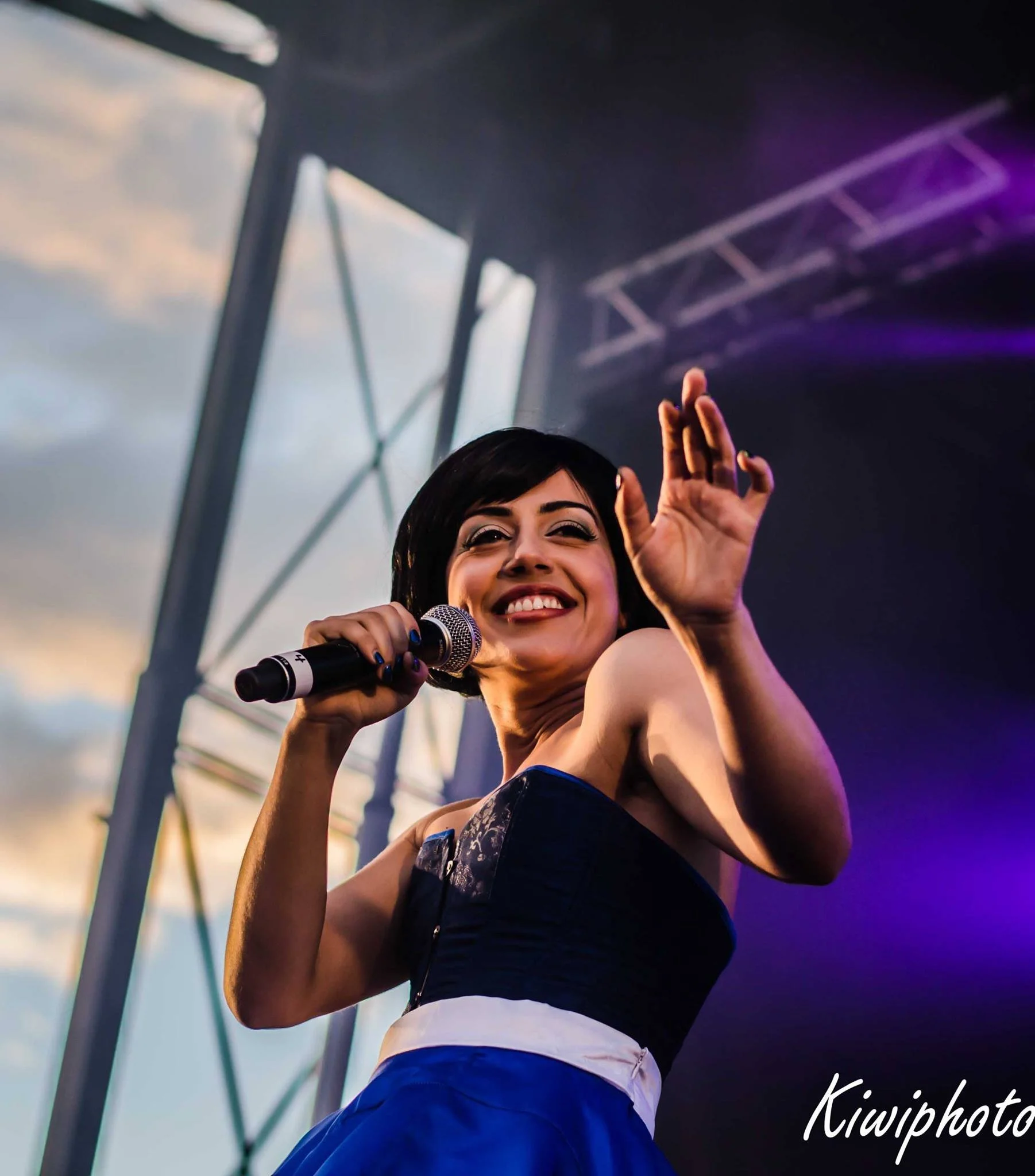 Une femme chante en performance lors d'un concert en extérieur, tenant un microphone et souriant.