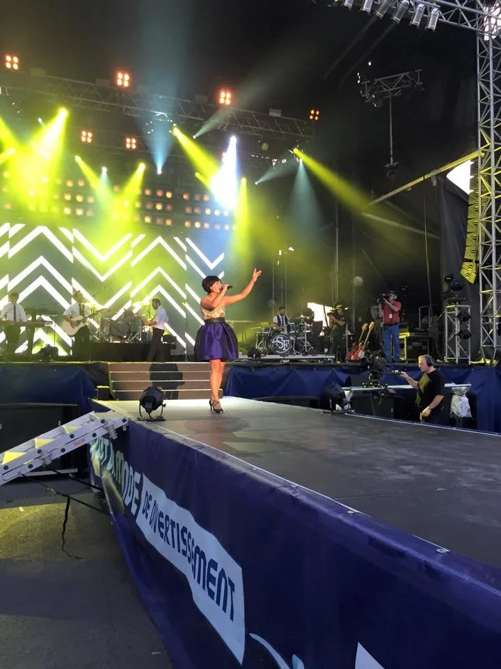 Une chanteuse en robe violette sur scène avec un groupe de musiciens, dans un festival de musique avec des éclairages colorés.