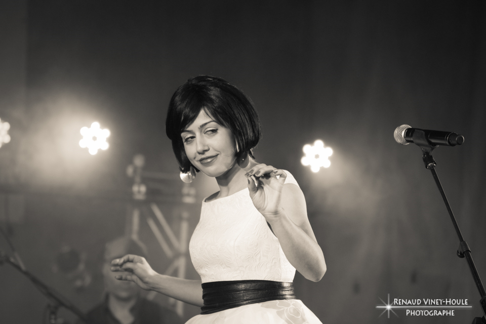 Une femme en noir et blanc sur scène, vêtue d'une robe blanche avec une ceinture noire, portant une coiffure courte et coiffée, se tenant près d'un microphone lors d'une performance musicale.