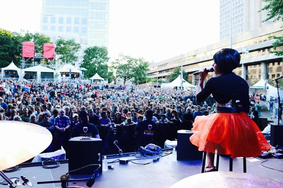 Une chanteuse en noir et orange performe devant une grande foule dans un événement en plein air, avec des tentes blanches et des bâtiments modernes en arrière-plan.
