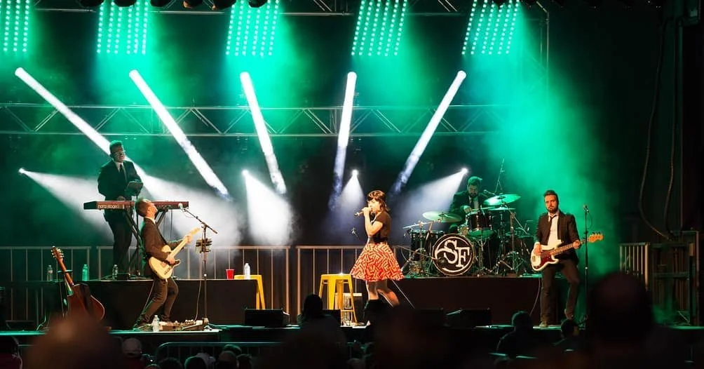 Un groupe de musiciens en concert sur scène avec éclairage vert et blanc, une femme chanteuse au centre, accompagnée de guitaristes et d'un batteur.