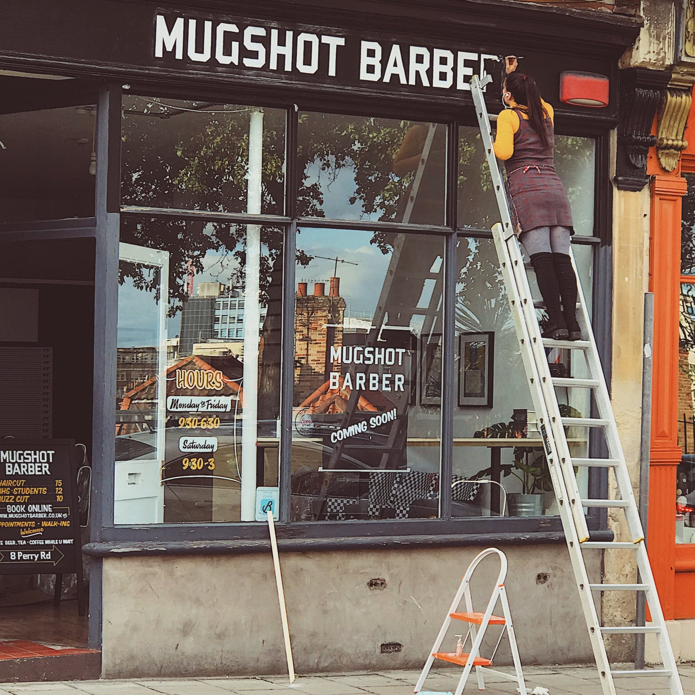 Photo of female signwriter up a large ladder hand-painting a shopfront.