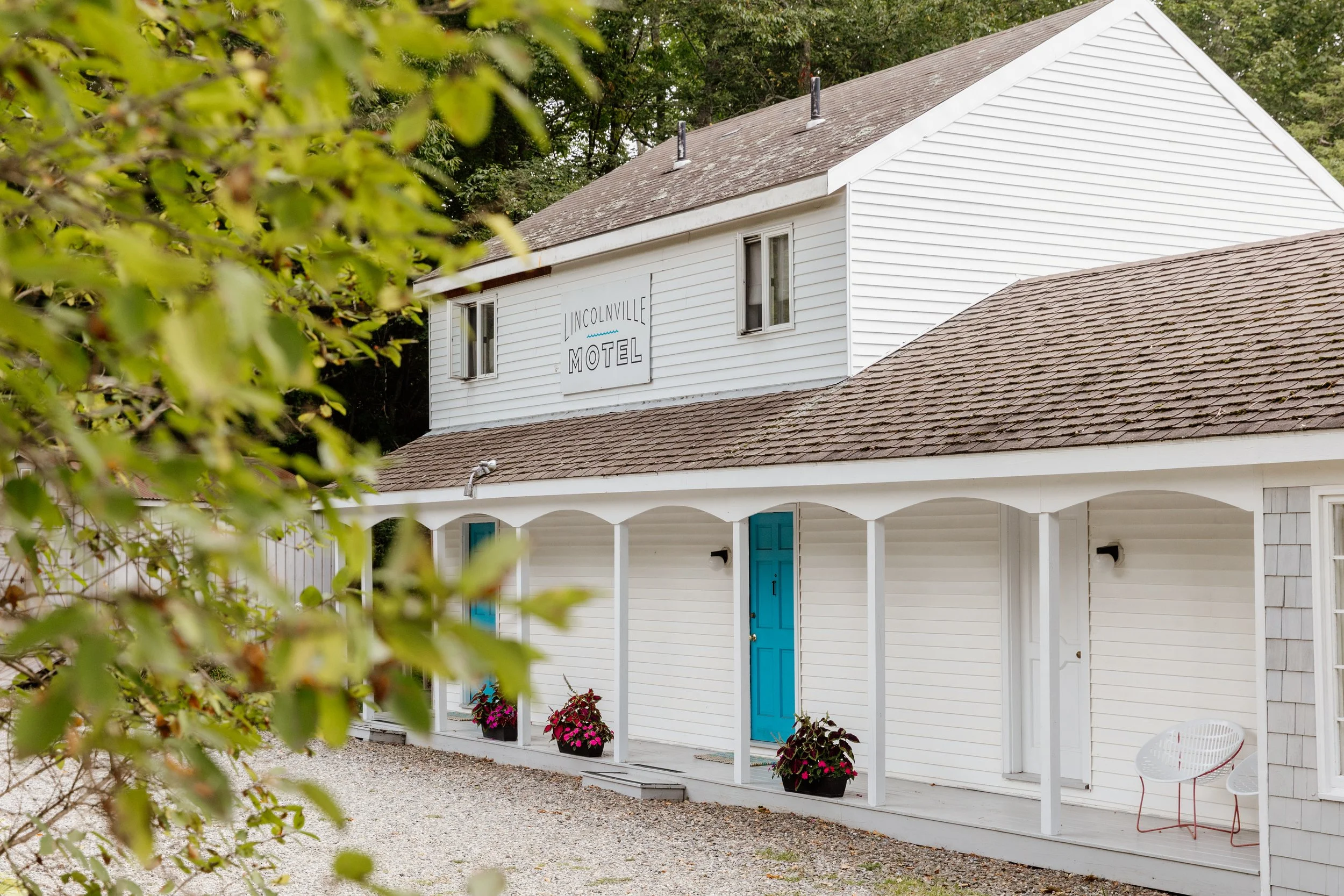 The welcoming exterior of the Lincolnville Motel in Lincolnville, Maine, a white two-story building with blue doors, a wraparound porch, potted flowers, and a gravel pathway nestled among the trees.