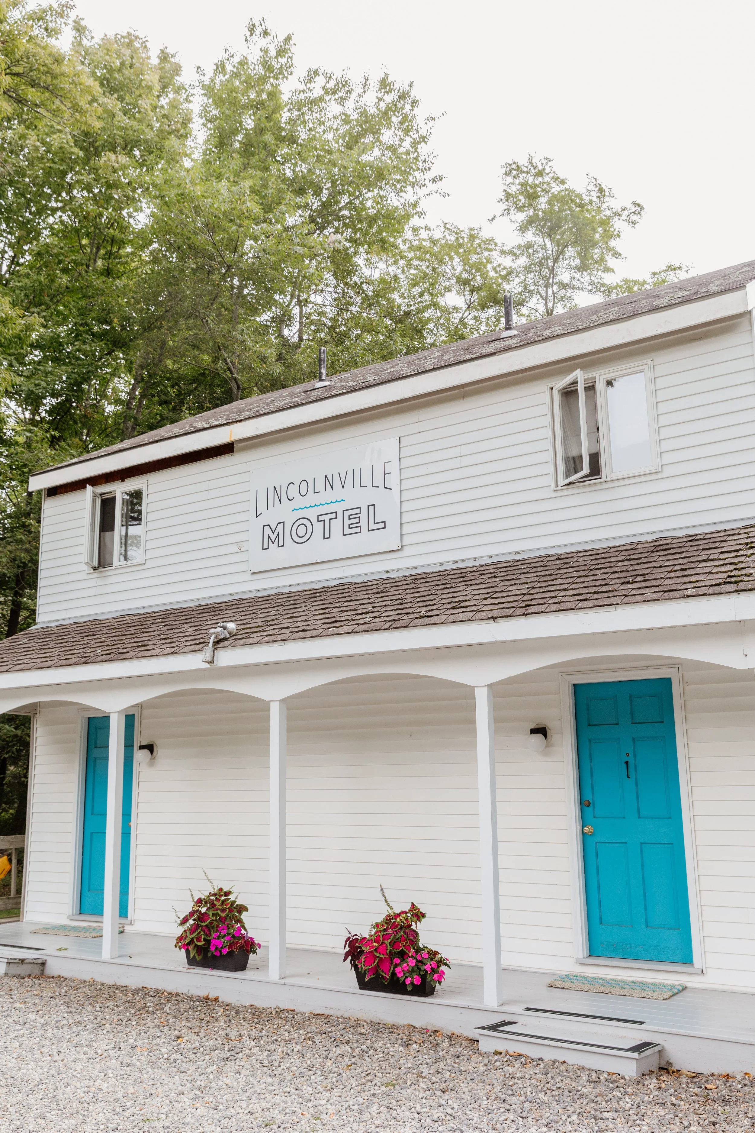 White two-story building with blue doors and a sign that reads 'Lincolnville Motel'. The building has a small porch with two flower pots containing pink and red flowers, and is set against a background of green trees.