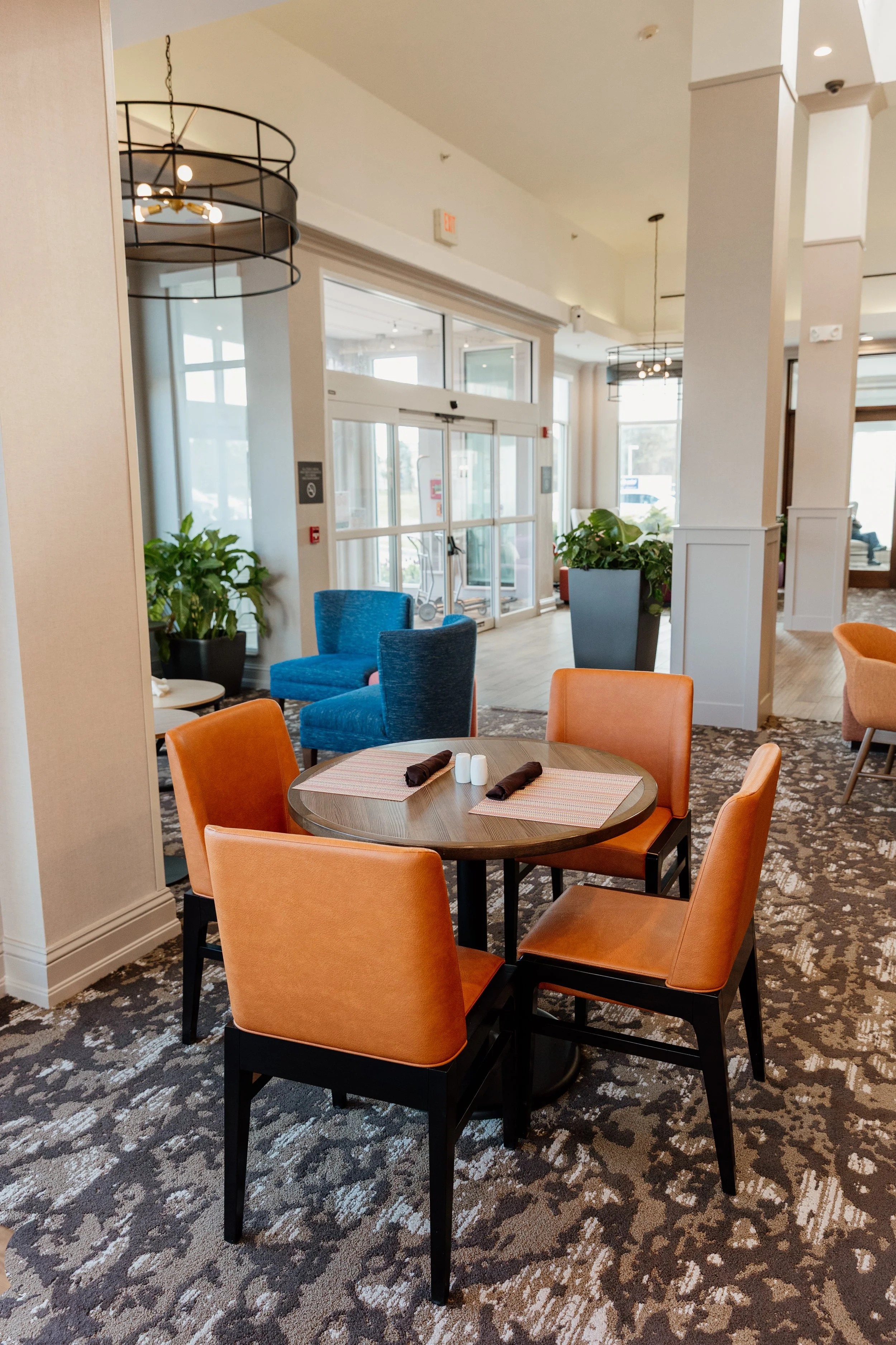 A round dining table set with four orange chairs, place settings with rolled napkins, salt and pepper shakers in a modern, well-lit restaurant or cafe.