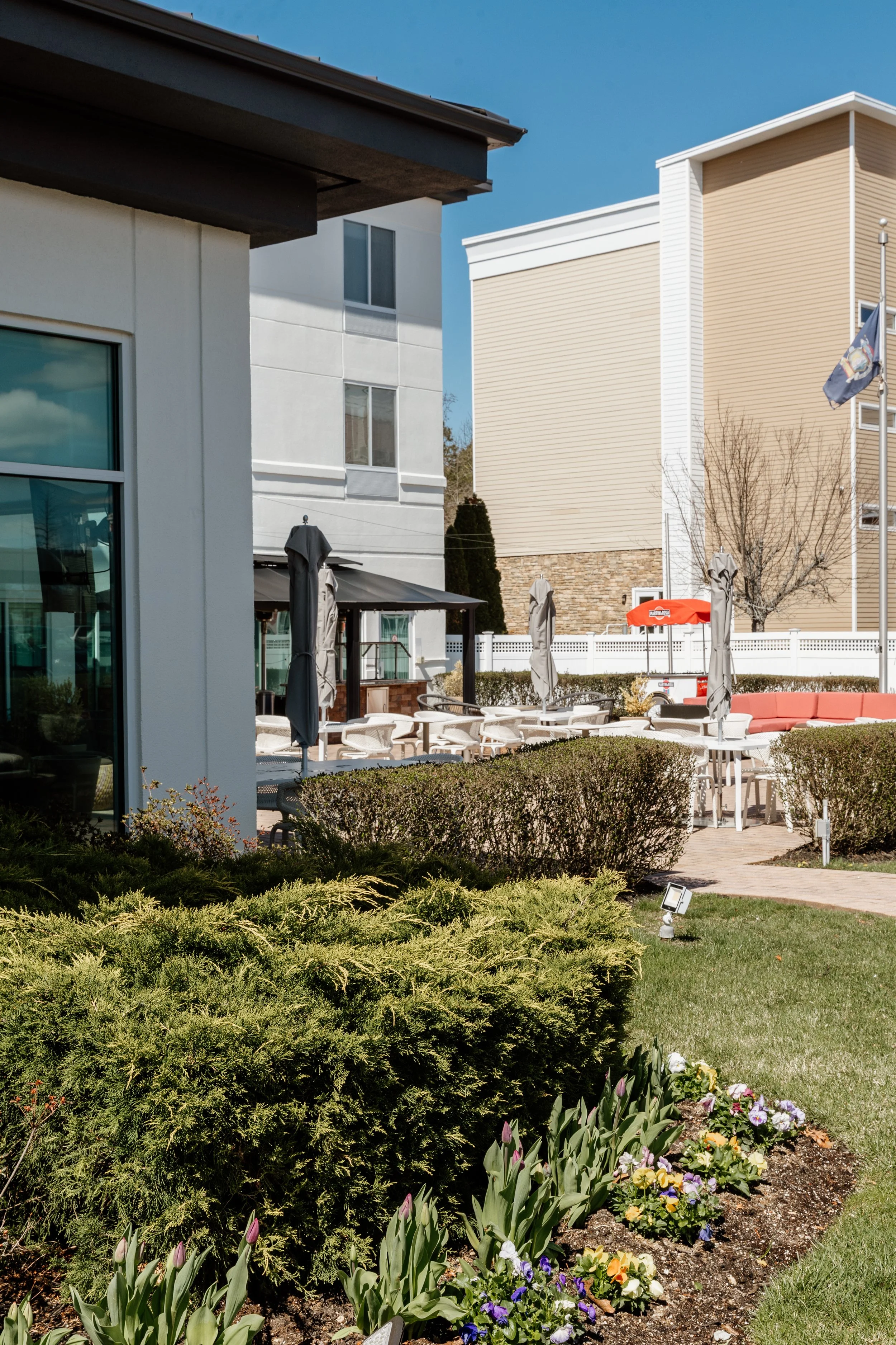 Outdoor patio area with tables, chairs, and umbrellas, surrounded by bushes and flower beds, with multi-story residential buildings in the background and a flagpole with a flag.