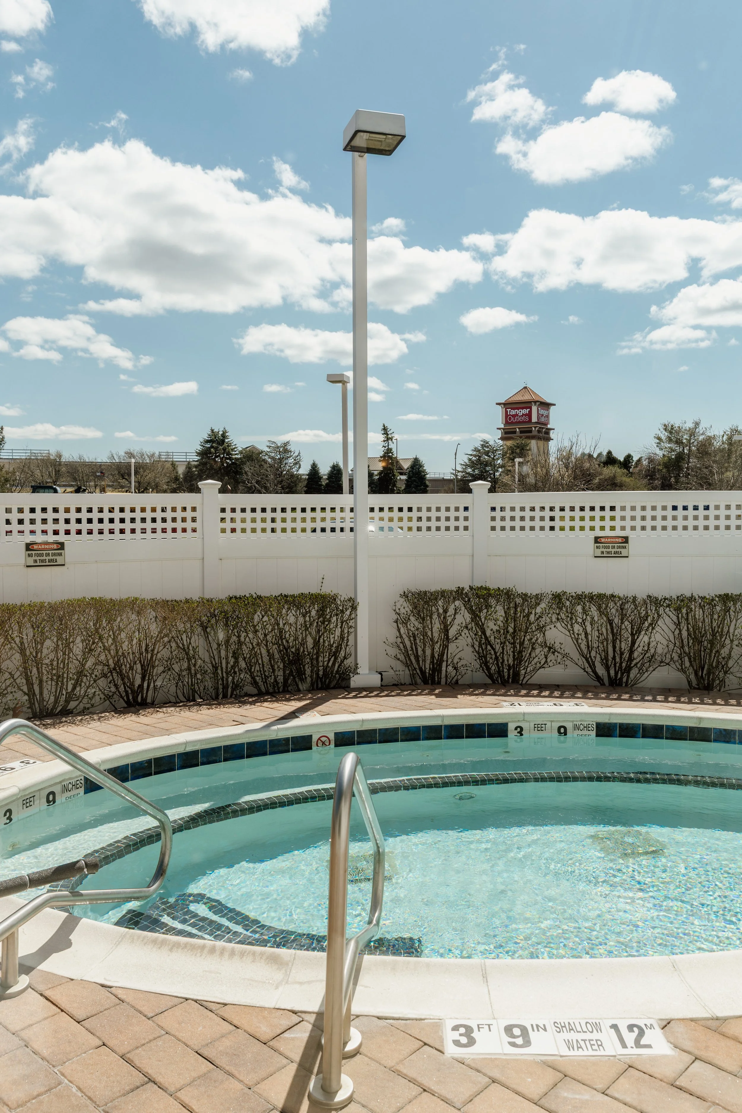 Empty outdoor swimming pool with a white fence, bushes, and a blue sky with clouds.