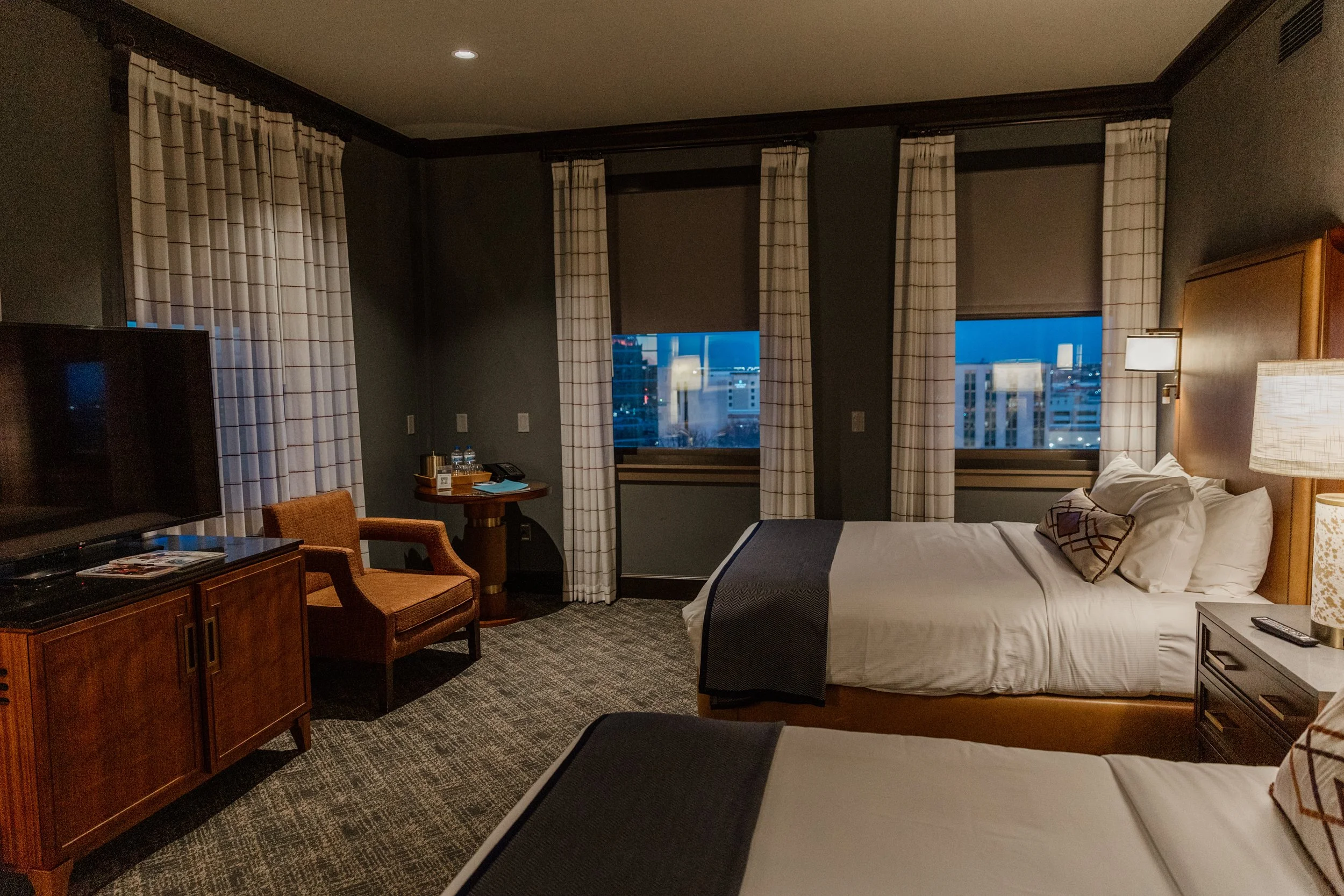 A hotel room with two beds, a chair, a TV, and a window with city view at dusk.