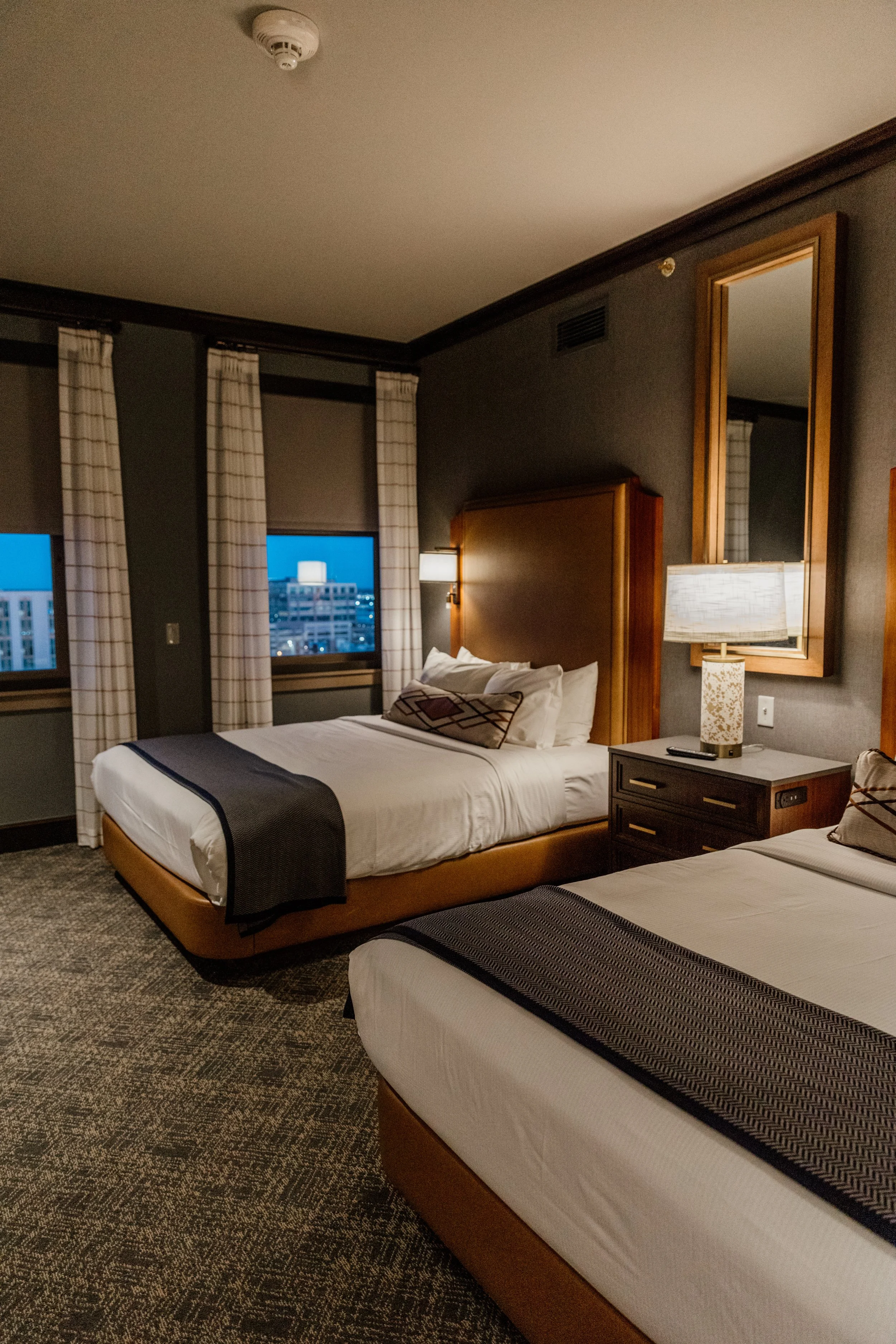 A hotel room with two beds, nightstand, lamp, windows with curtains, and city view outside at dusk.