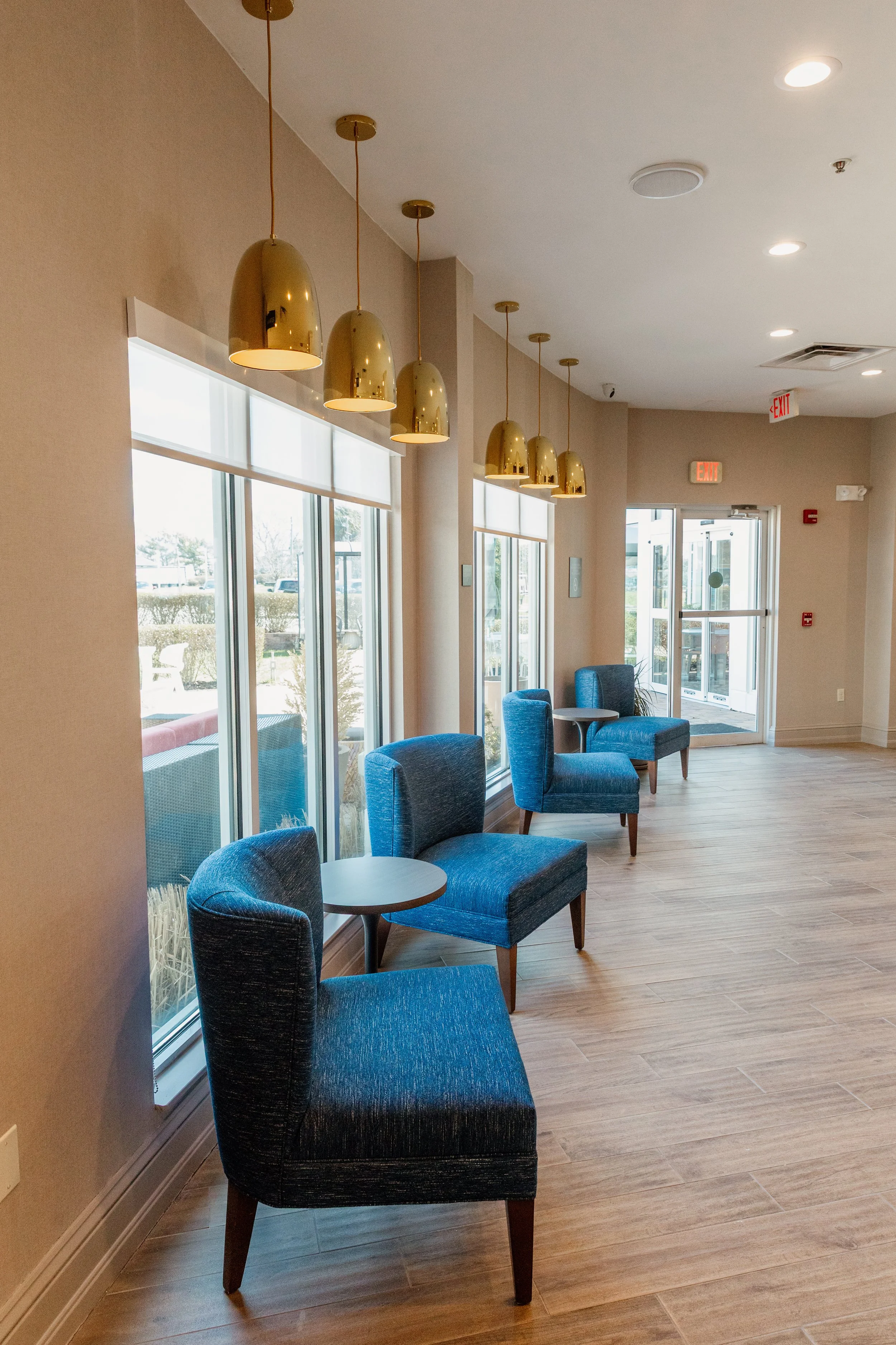 A brightly lit indoor space with large windows, blue upholstered chairs, small round tables, hanging gold pendant lights, and double glass doors leading outside.
