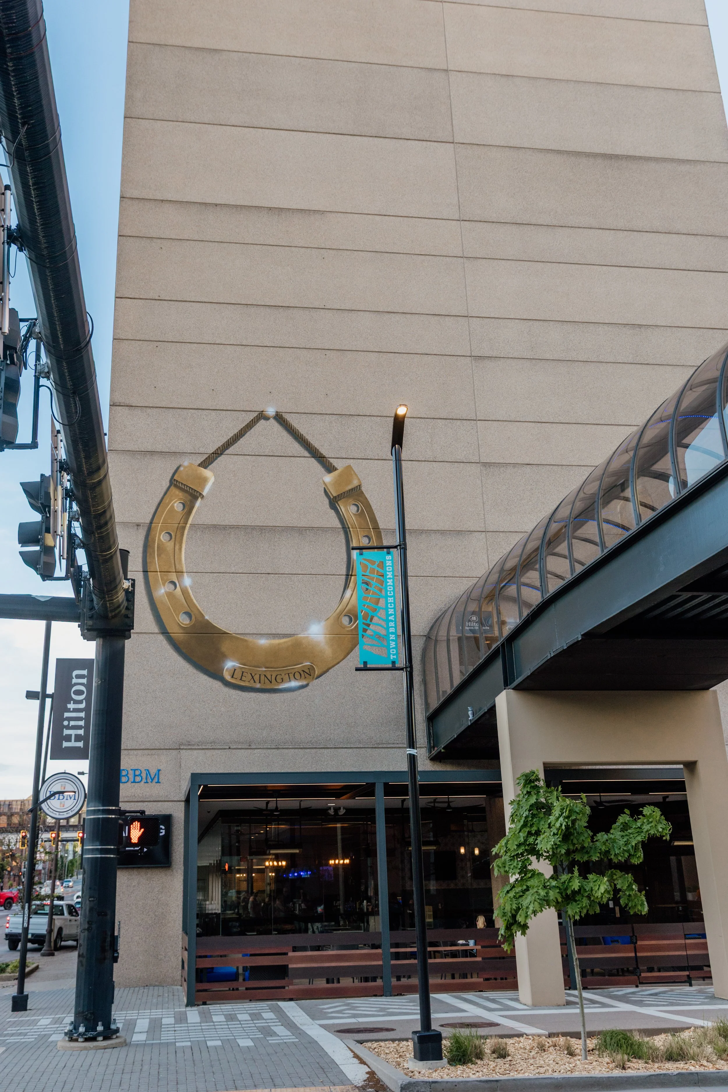 The exterior of a building with a large horseshoe-shaped sign reading 'LEXINGTON' and a blue banner for the Kentucky Derby, with street elements like traffic lights, a sidewalk, and a small tree.
