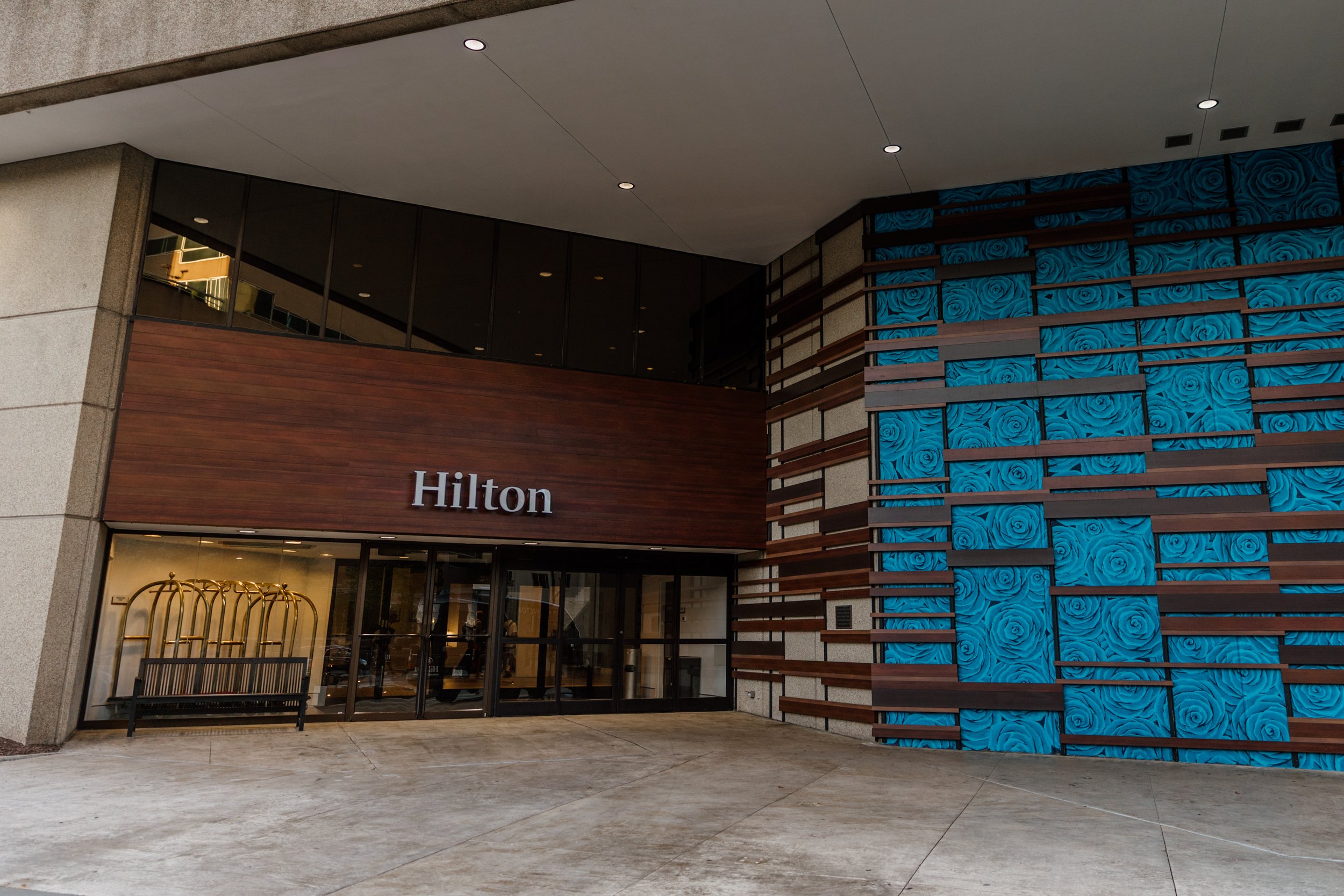 Exterior view of the Hilton hotel entrance with a wooden sign, glass doors, luggage cart, and decorative blue floral wall panels on the right.