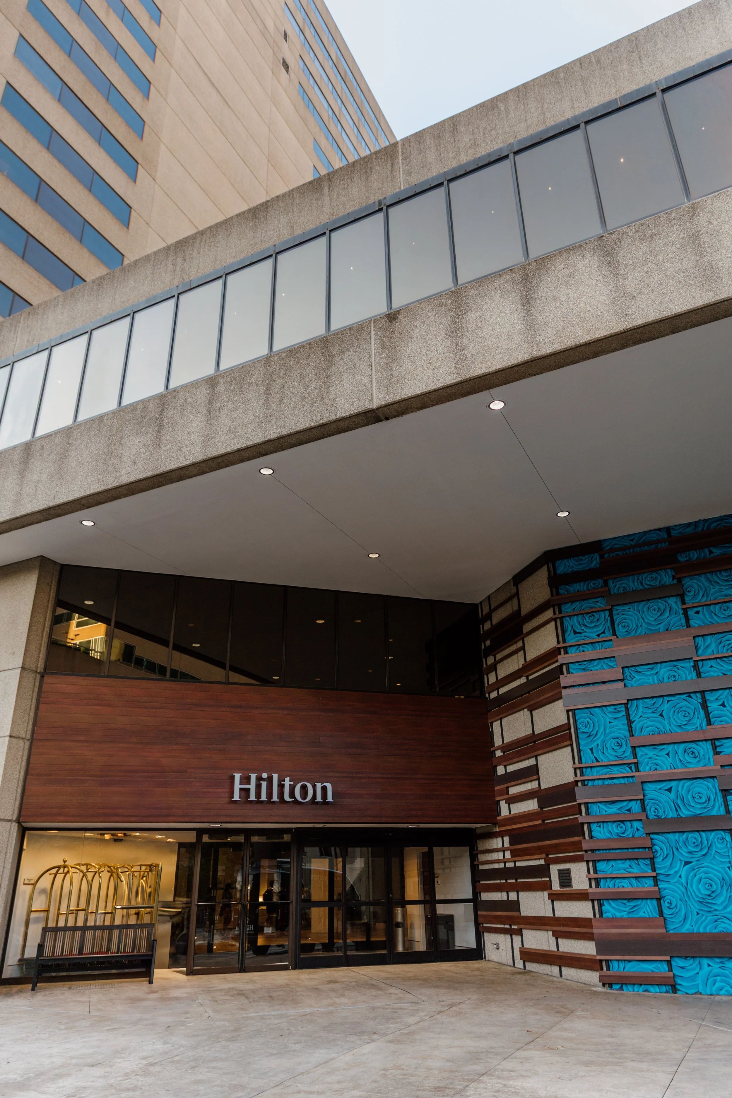 Exterior view of the Hilton hotel entrance with wooden signage, glass doors, and modern architectural design including patterned blue panels.