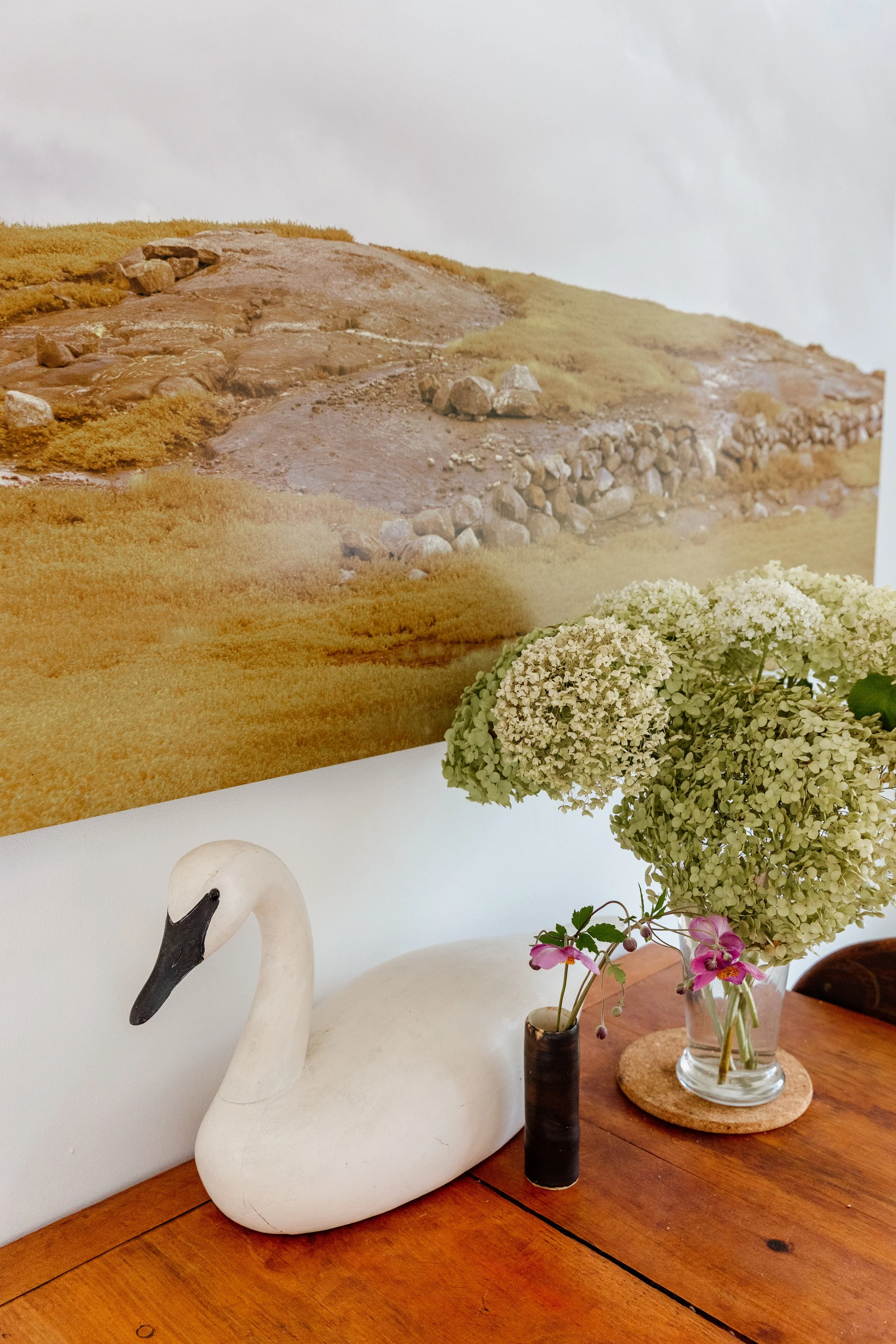 Decorative scene with a white swan figurine, two vases with flowers, and a large picture of a rocky hillside landscape in the background.