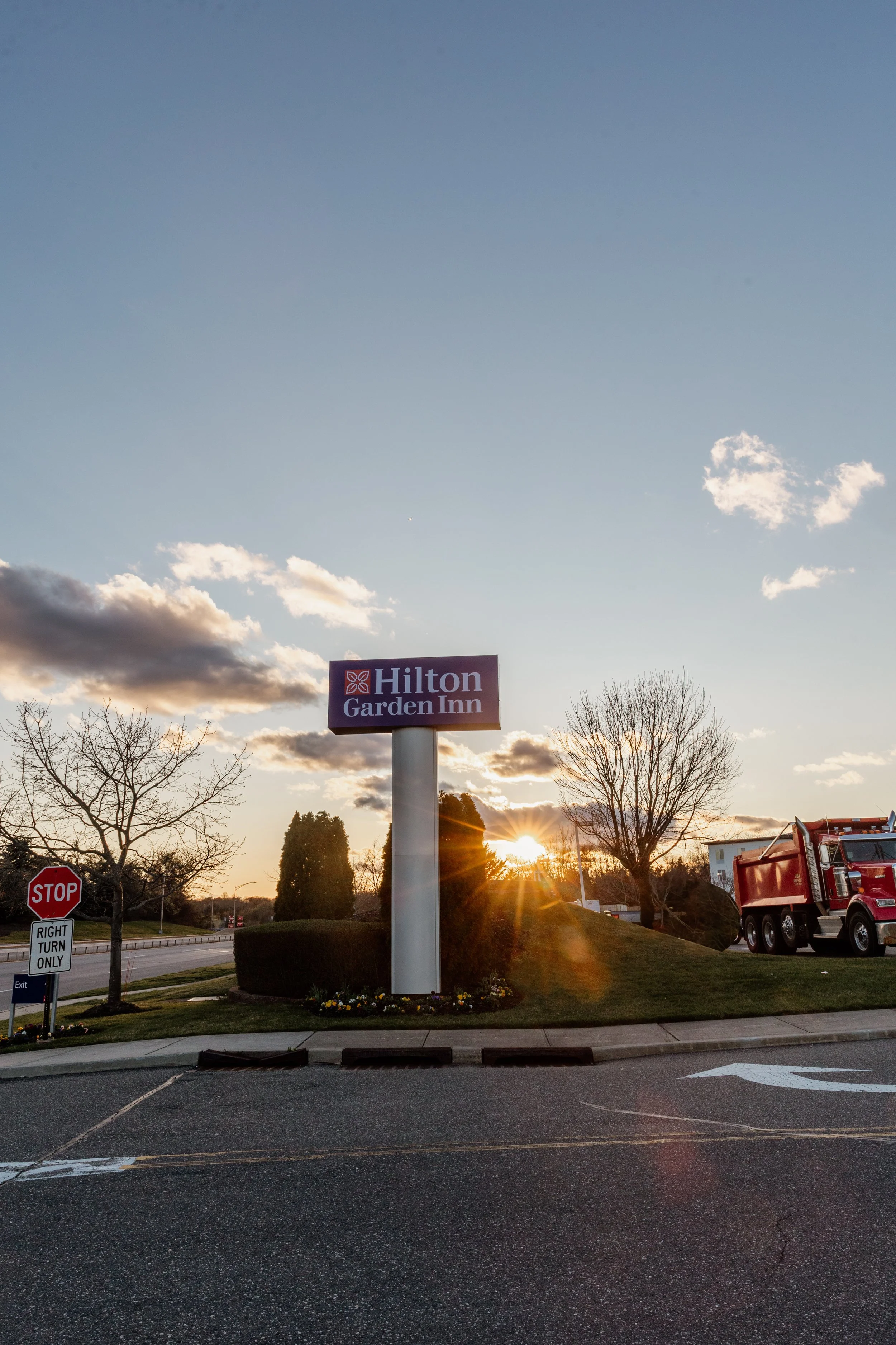 Sunset behind a Hilton Garden Inn sign surrounded by trees in a parking lot.