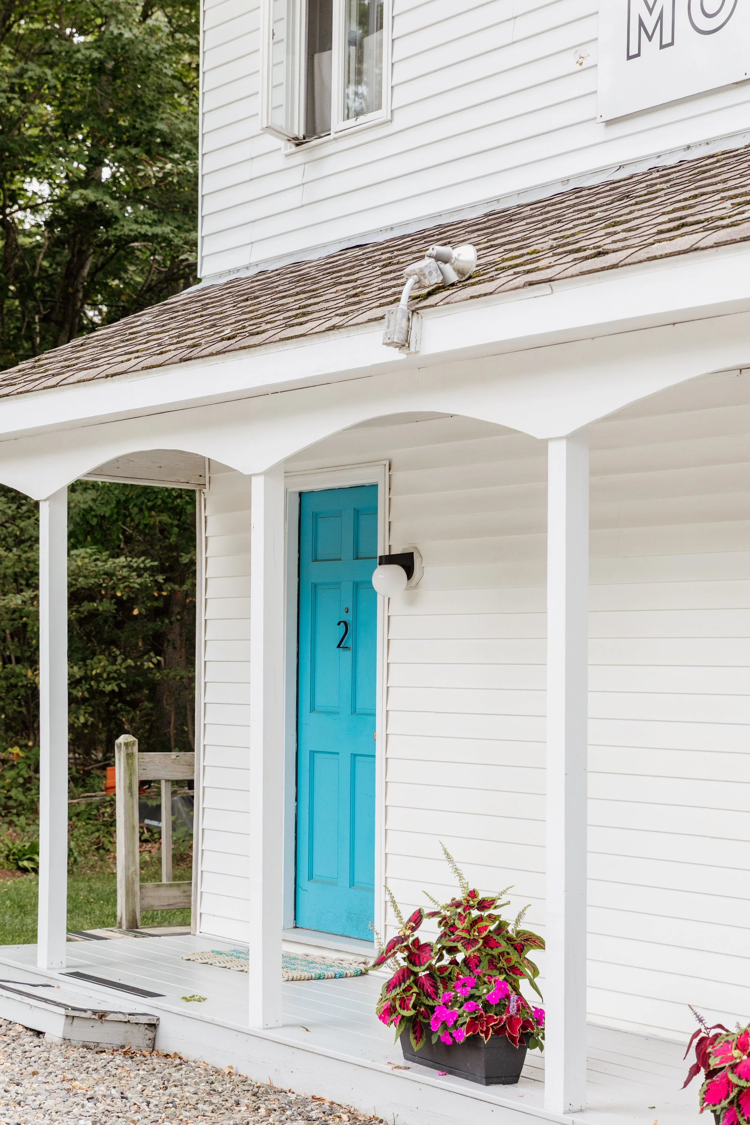 Front entrance of a white house with a blue door, a potted plant with pink and green flowers, and a white porch with a partial railing.
