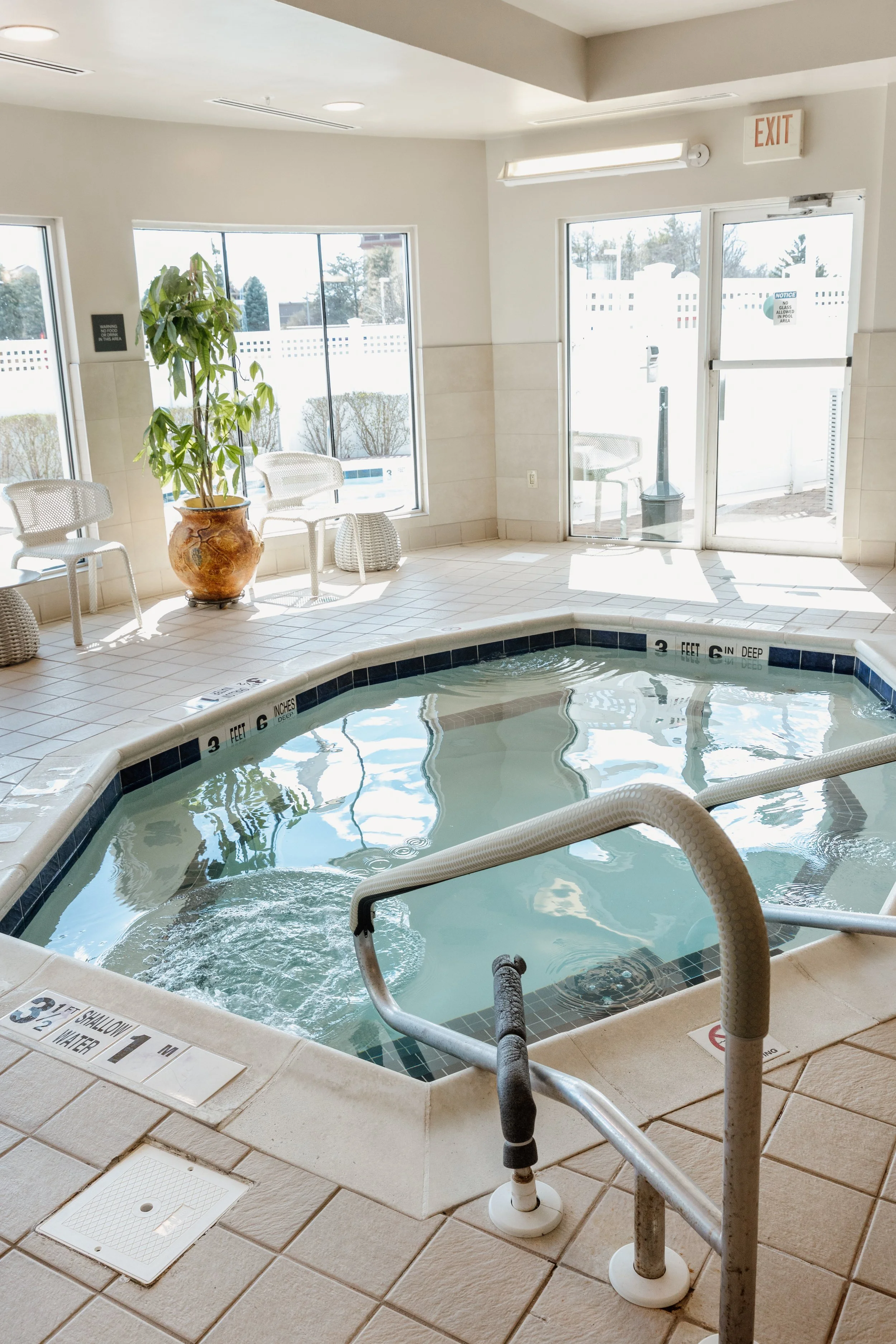 Indoor pool area with a small pool, white tiled floor, and large windows allowing sunlight. There are chairs, a potted plant, and an exit door visible.