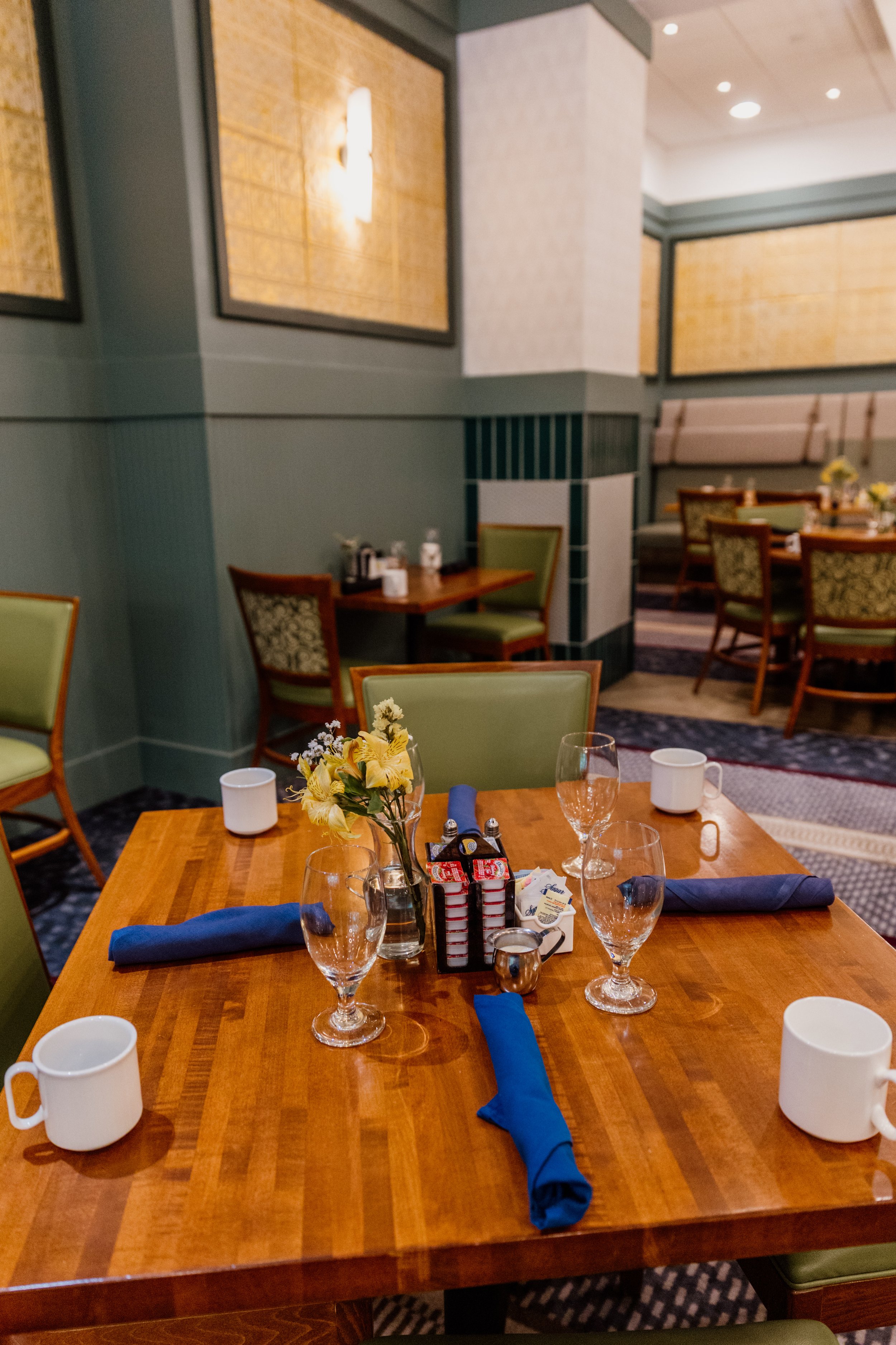 A table set for dining with four glasses, four white mugs, blue napkins, a small flower arrangement, and condiment bottles in a dining area with green chairs and wall decor.