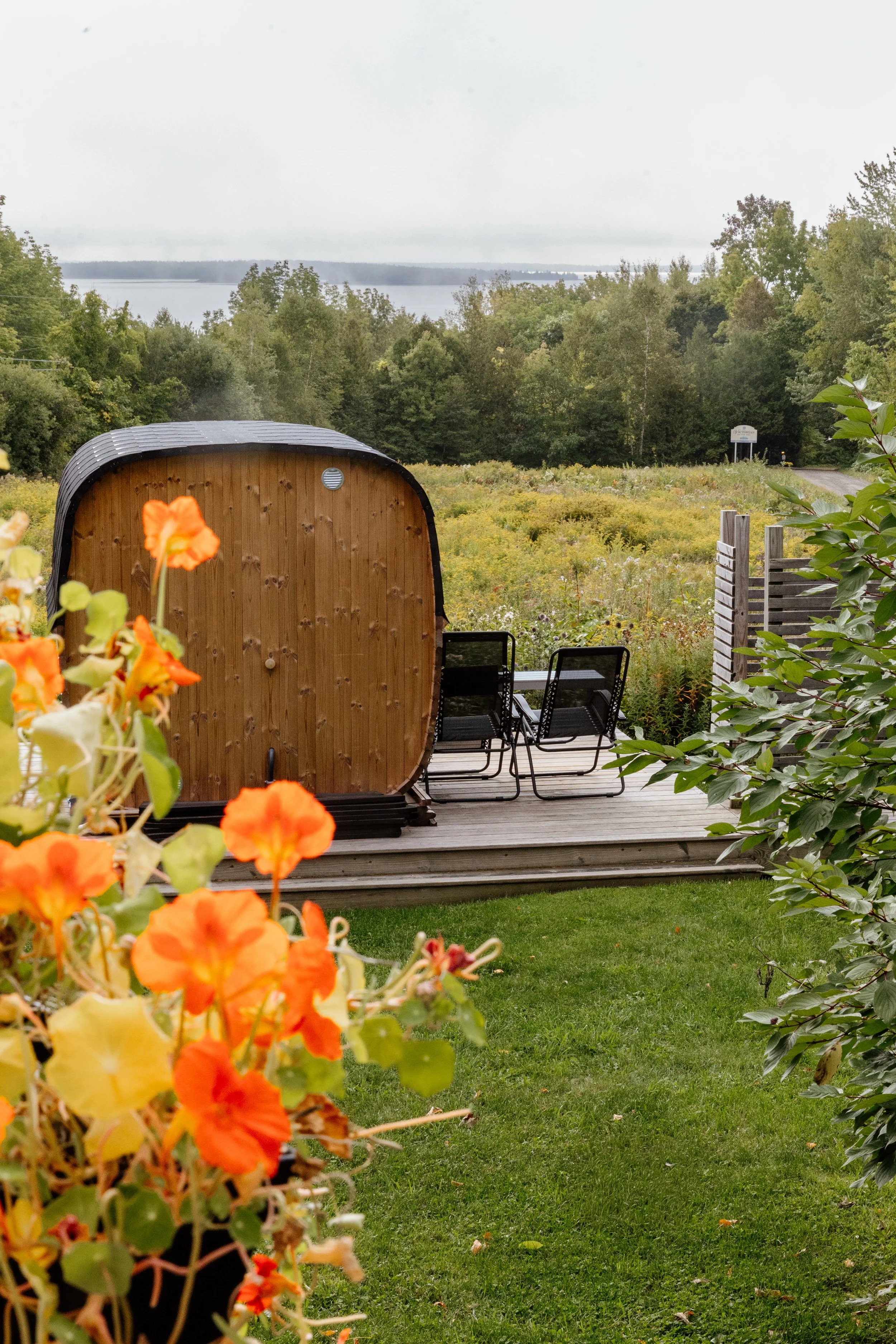A view of a backyard with a wooden deck, two black chairs, a garden shed with a curved roof, vibrant orange and yellow flowers in the foreground, and a body of water along with trees and an overcast sky in the background.