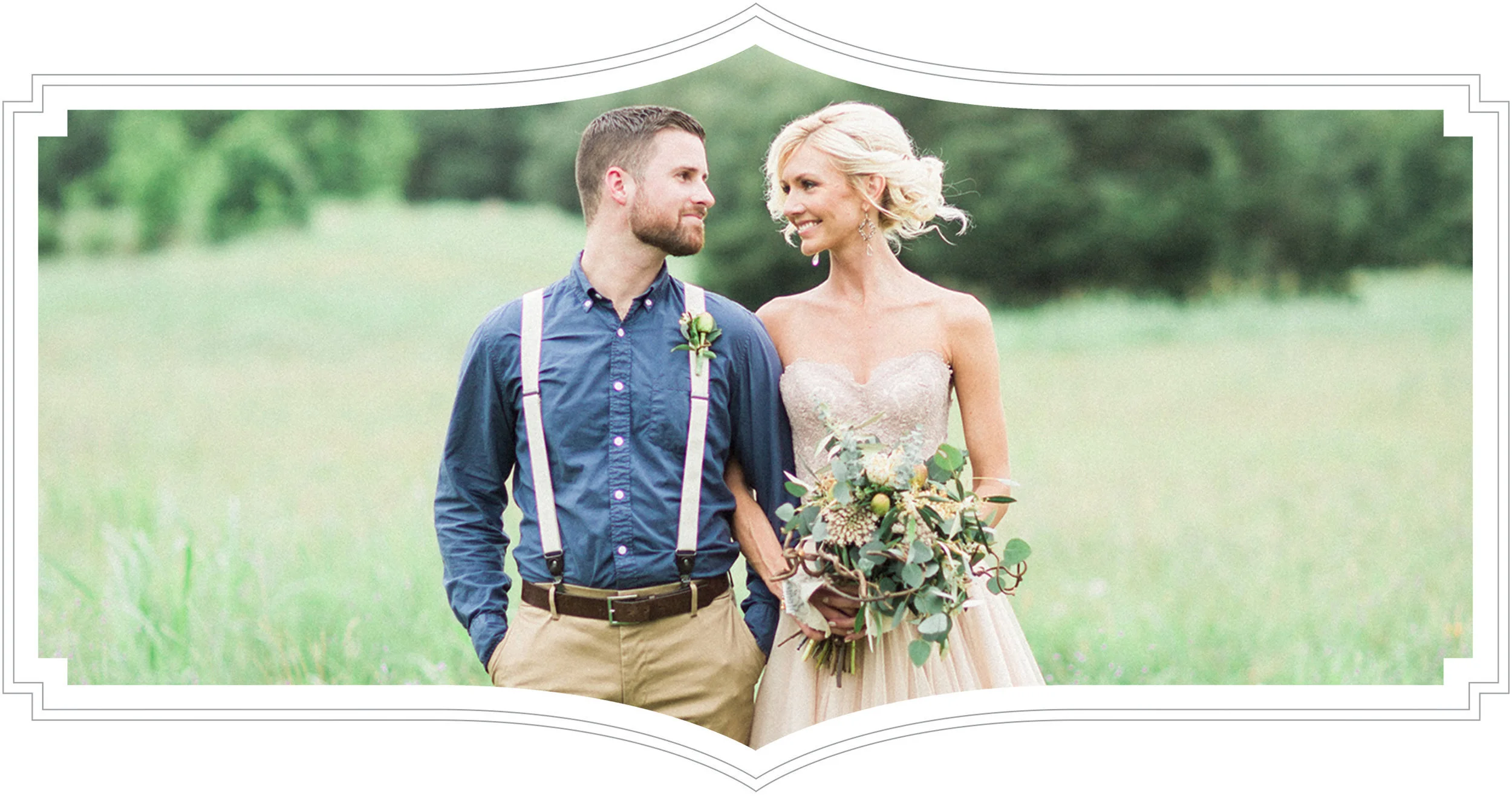 A bride and groom standing together in a field, looking at each other with smiles. The bride is holding a bouquet of flowers and greenery, wearing a strapless wedding dress. The groom is in a button-up shirt with suspenders, with hands in his pockets