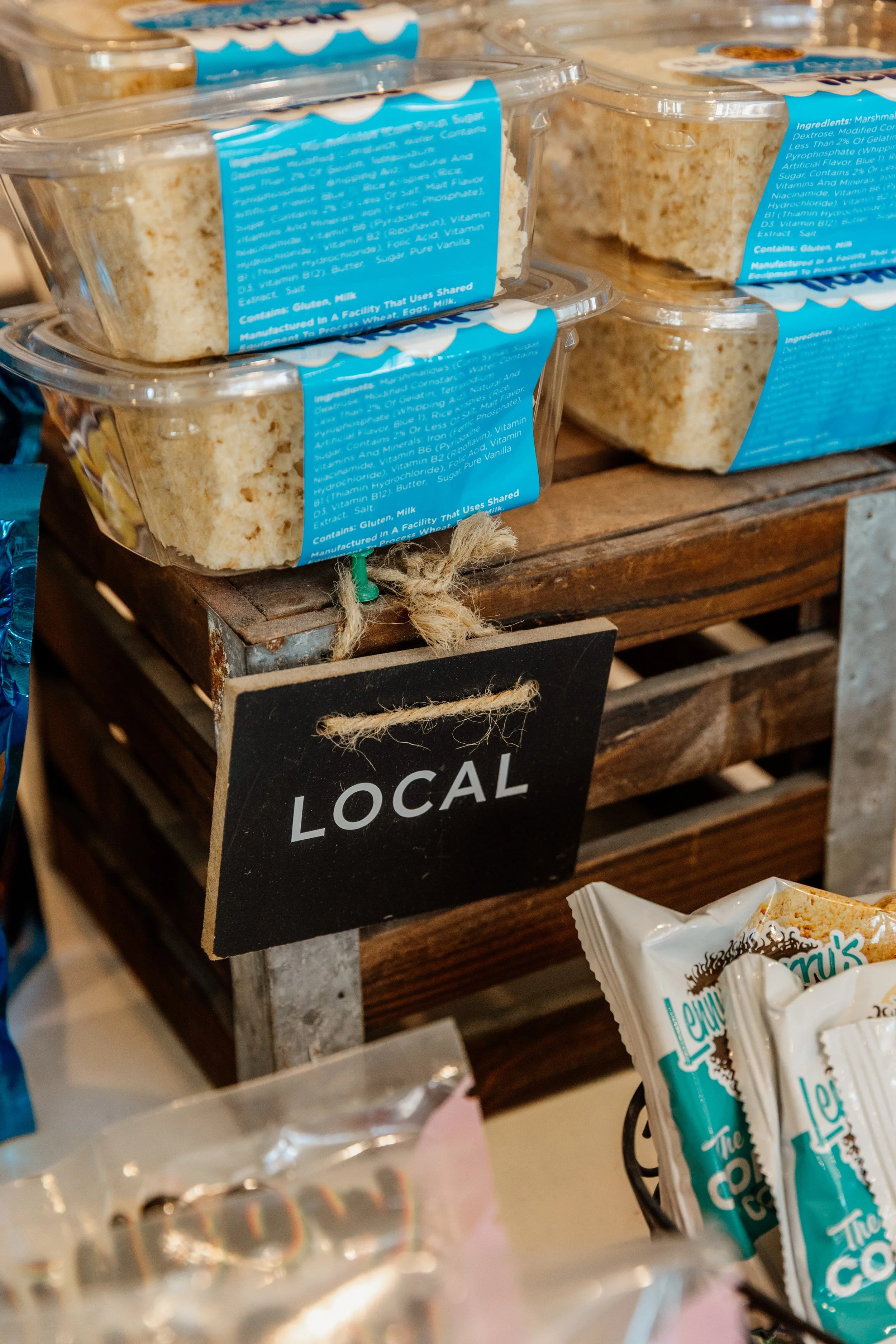 Clear plastic containers with slices of vanilla cake on display at a local market, with a black chalkboard sign labeled 'LOCAL' hanging below.