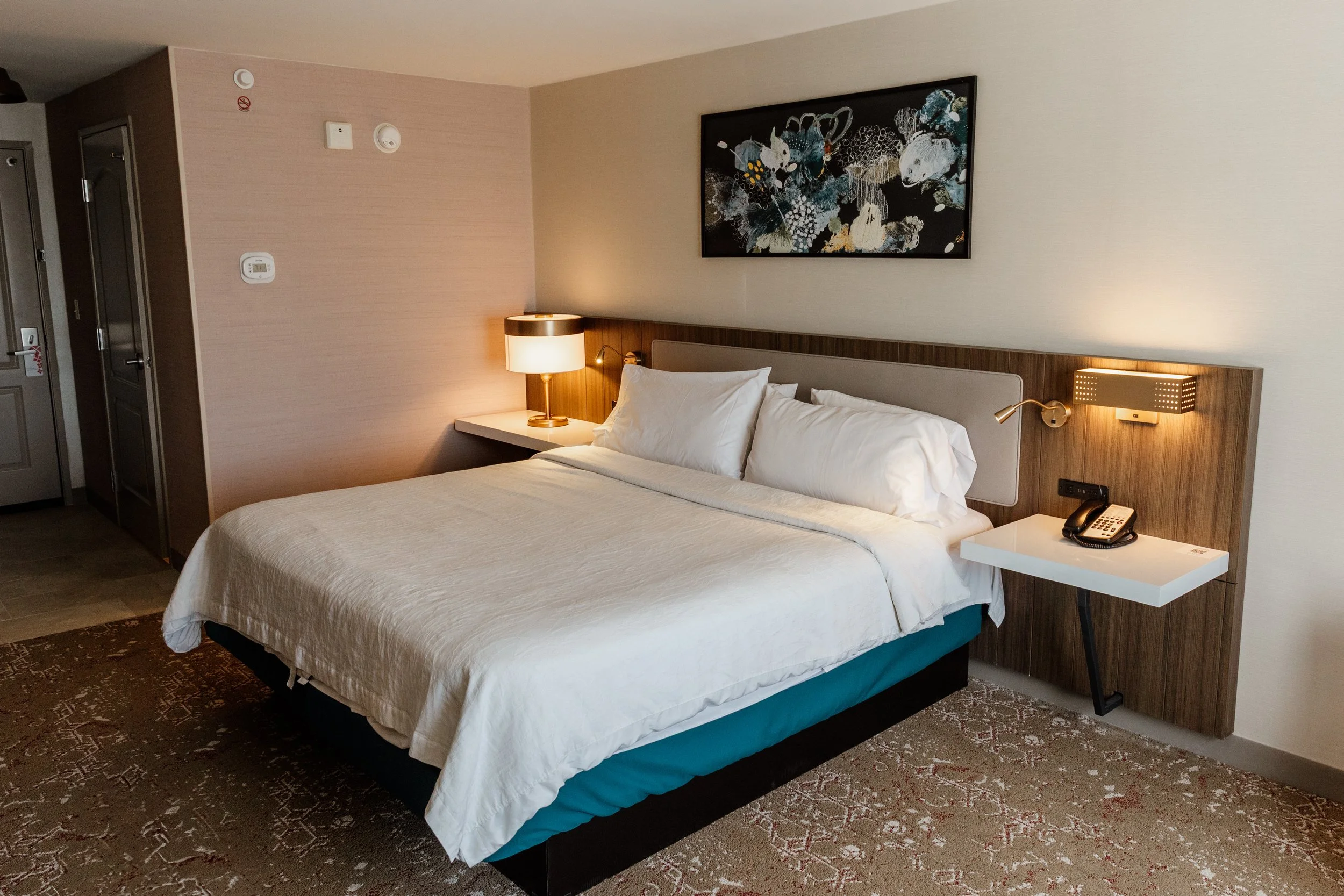 A crisp and well-appointed guest room at the Hilton Lexington Downtown in Lexington, Kentucky, with white bedding, warm wood tones, bedside lamps, and a framed abstract artwork above the bed.