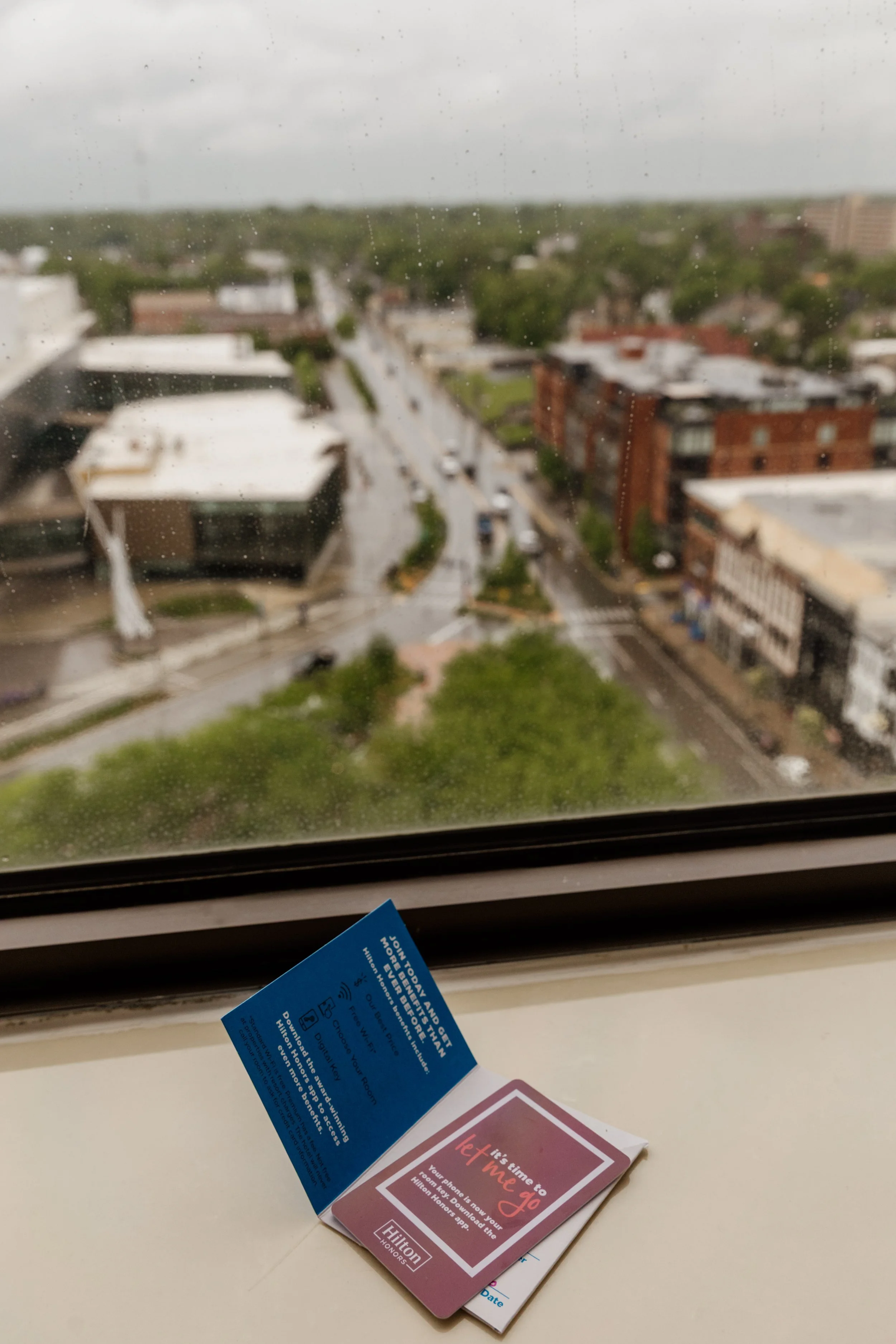 View of a rainy city street seen through a window with water droplets, with a Hilton hotel keycard and room information on the windowsill in the foreground.