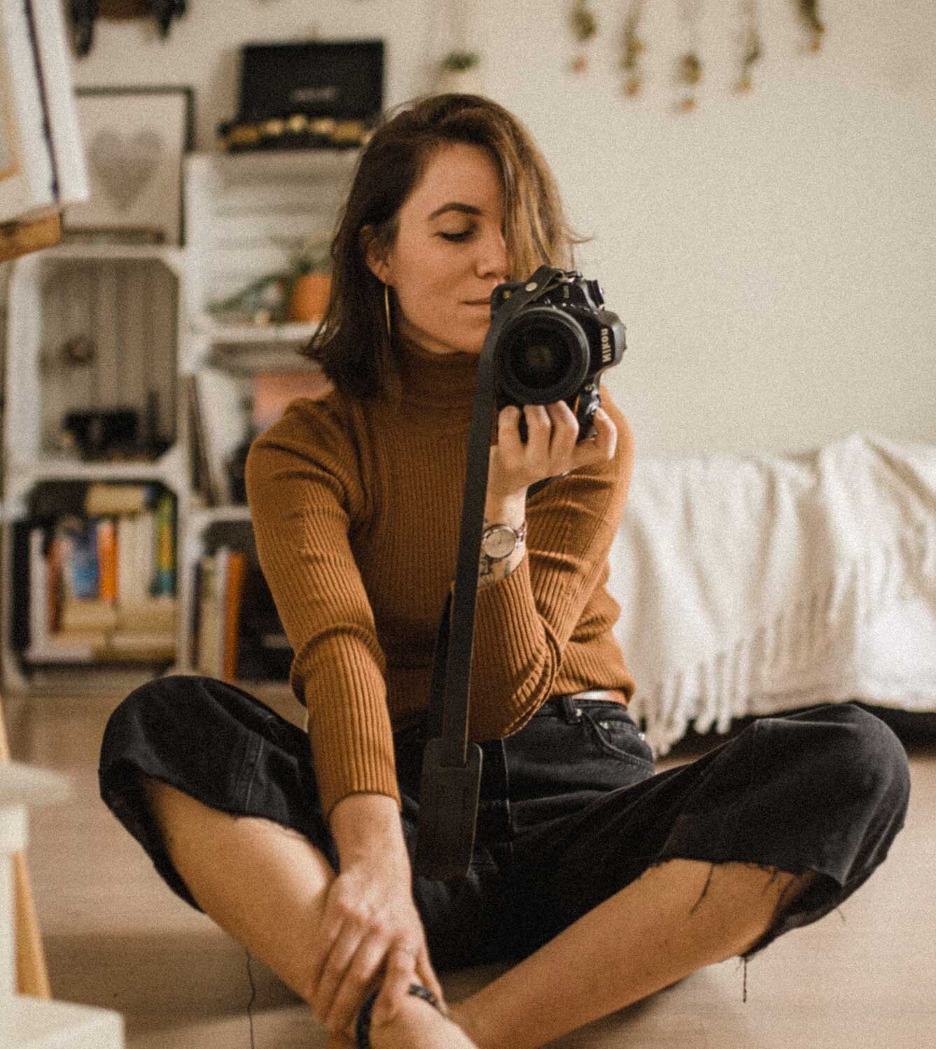 A woman with shoulder-length brown hair sitting cross-legged on the floor, taking a photo of herself in a mirror with a Canon camera. She is wearing a brown turtleneck, black ripped jeans, and a watch on her left wrist. The background features a white bookshelf filled with books and decorative items, and a bed with a white blanket.