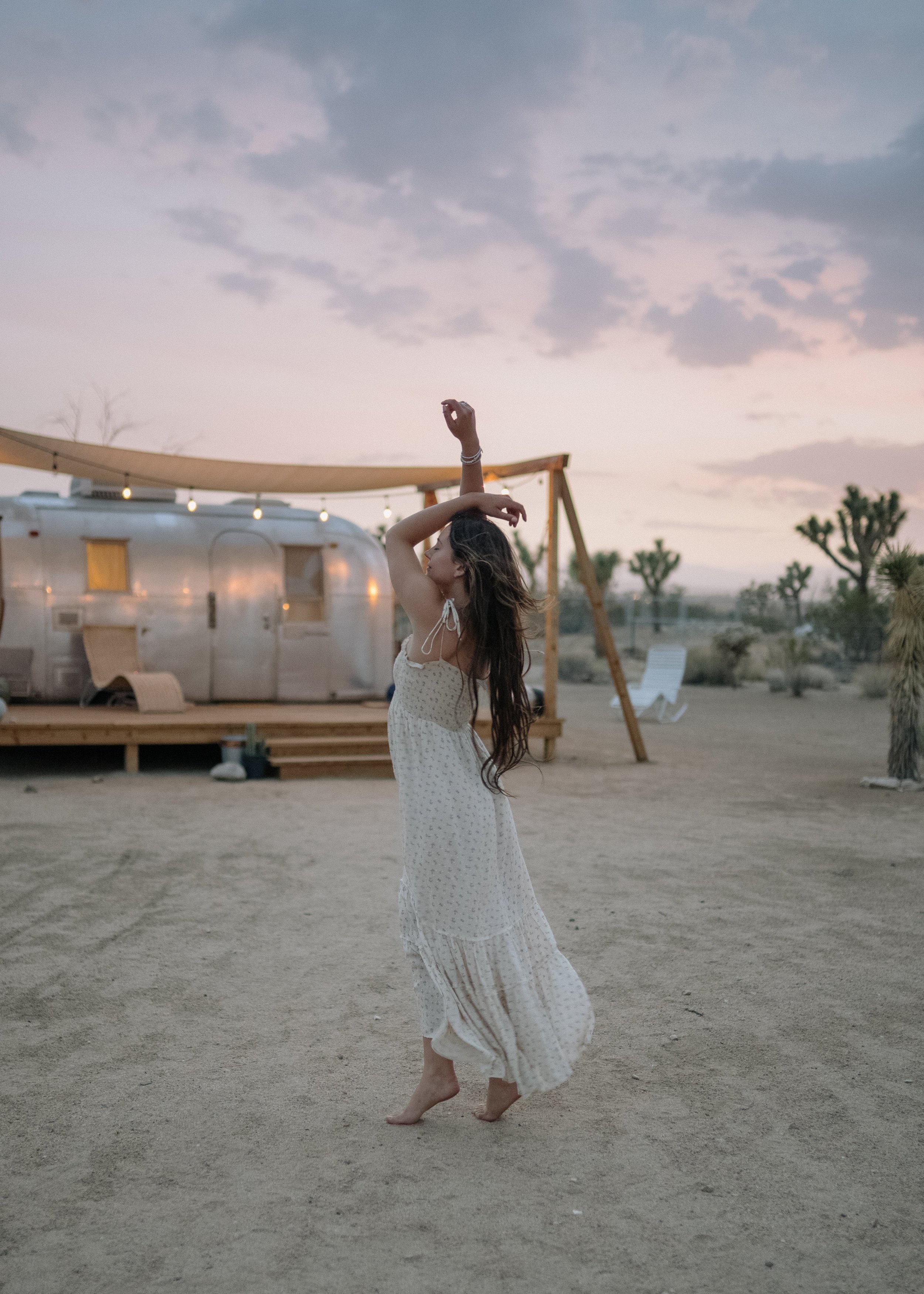 A woman dancing barefoot on a sandy desert at sunset, wearing a flowing white dress, with a vintage trailer and Joshua trees in the background.