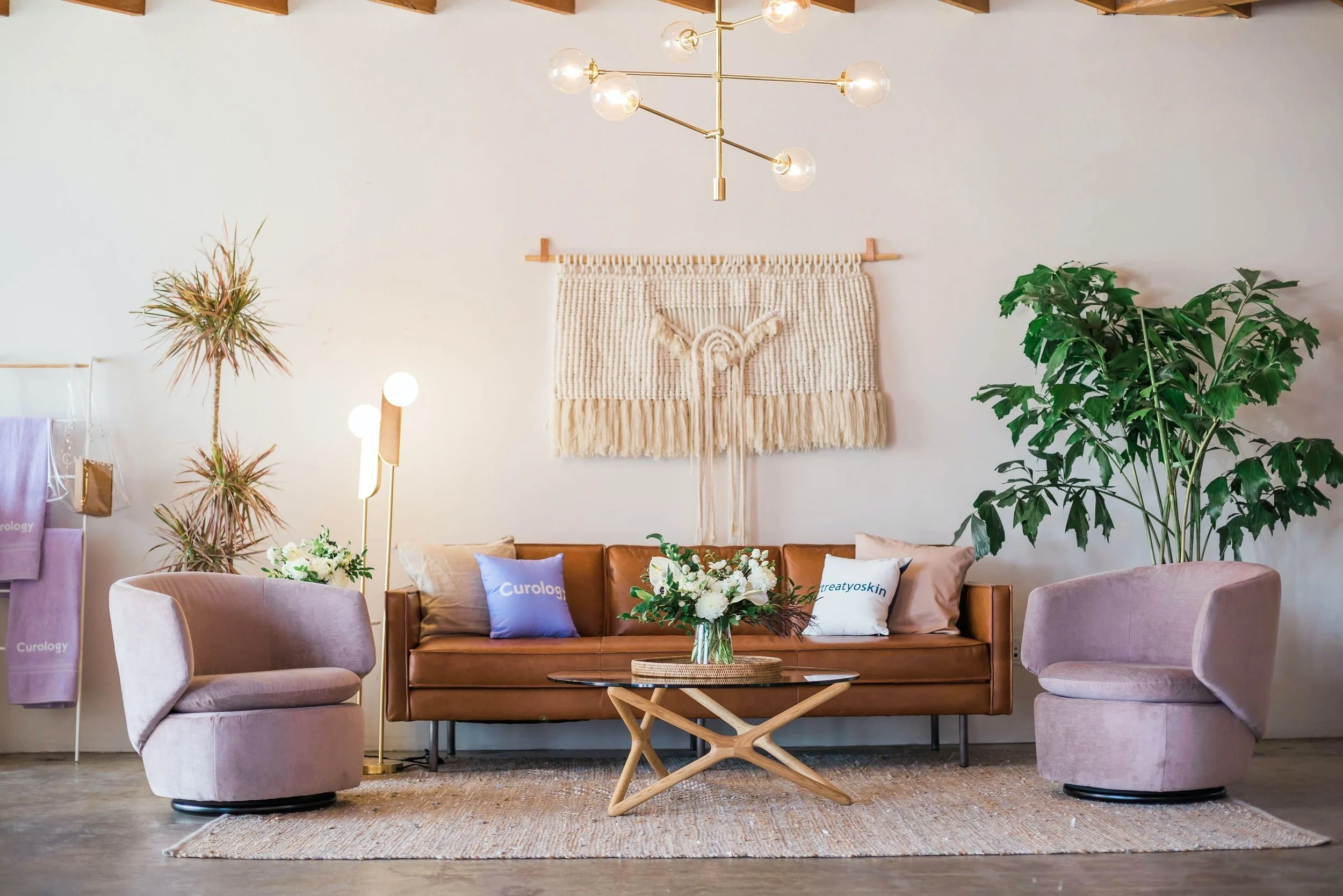 Living room with a leather sofa, pink swivel chairs, and a wooden coffee table with a flower vase. Decor includes indoor plants, wall hanging, and cushions with logos. Modern light fixtures and a woven rug complete the scene.