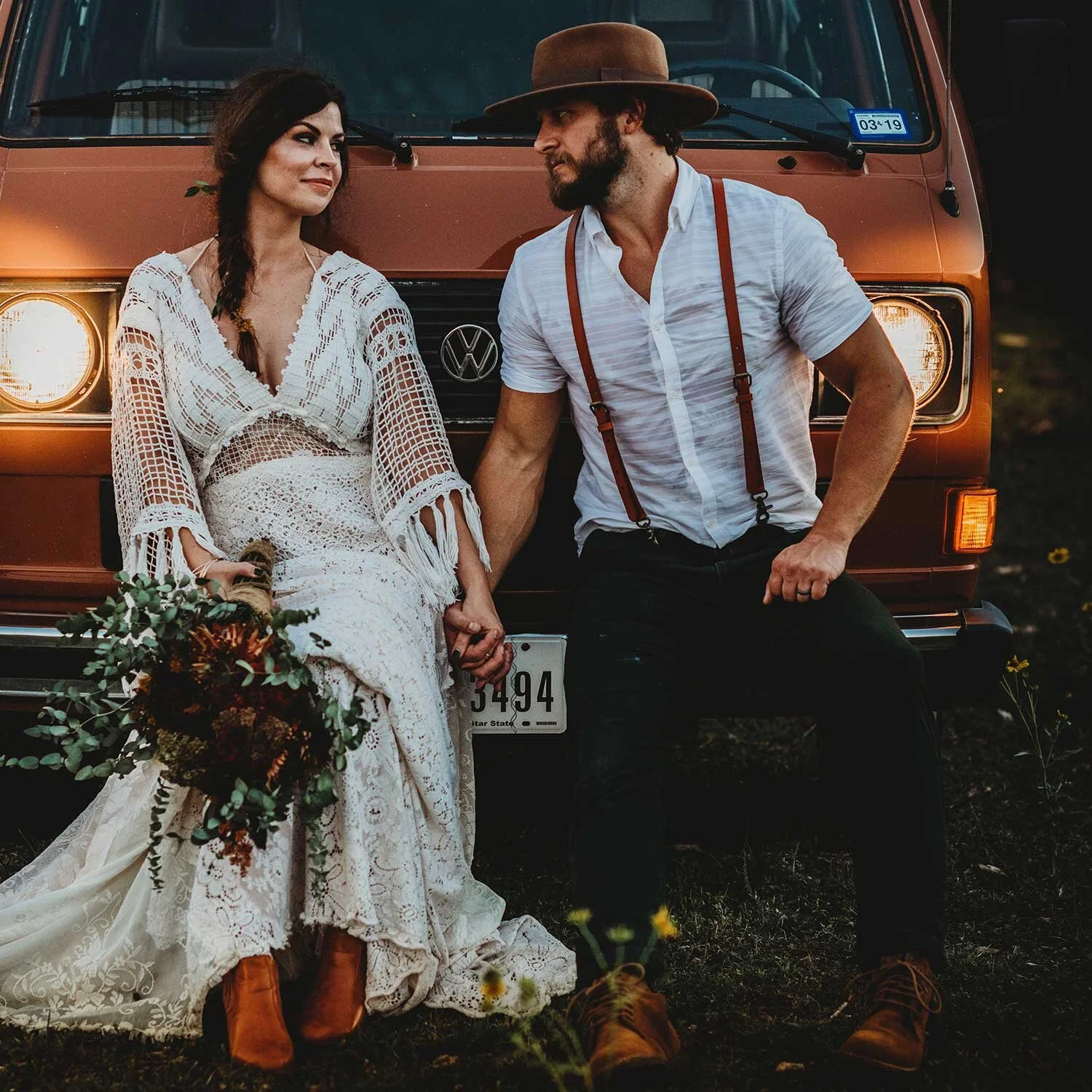 A couple sits in front of an orange vintage van, holding hands. The woman wears a white crochet dress and brown boots, holding a bouquet of flowers. The man wears a white shirt, brown hat, suspenders, and brown boots.