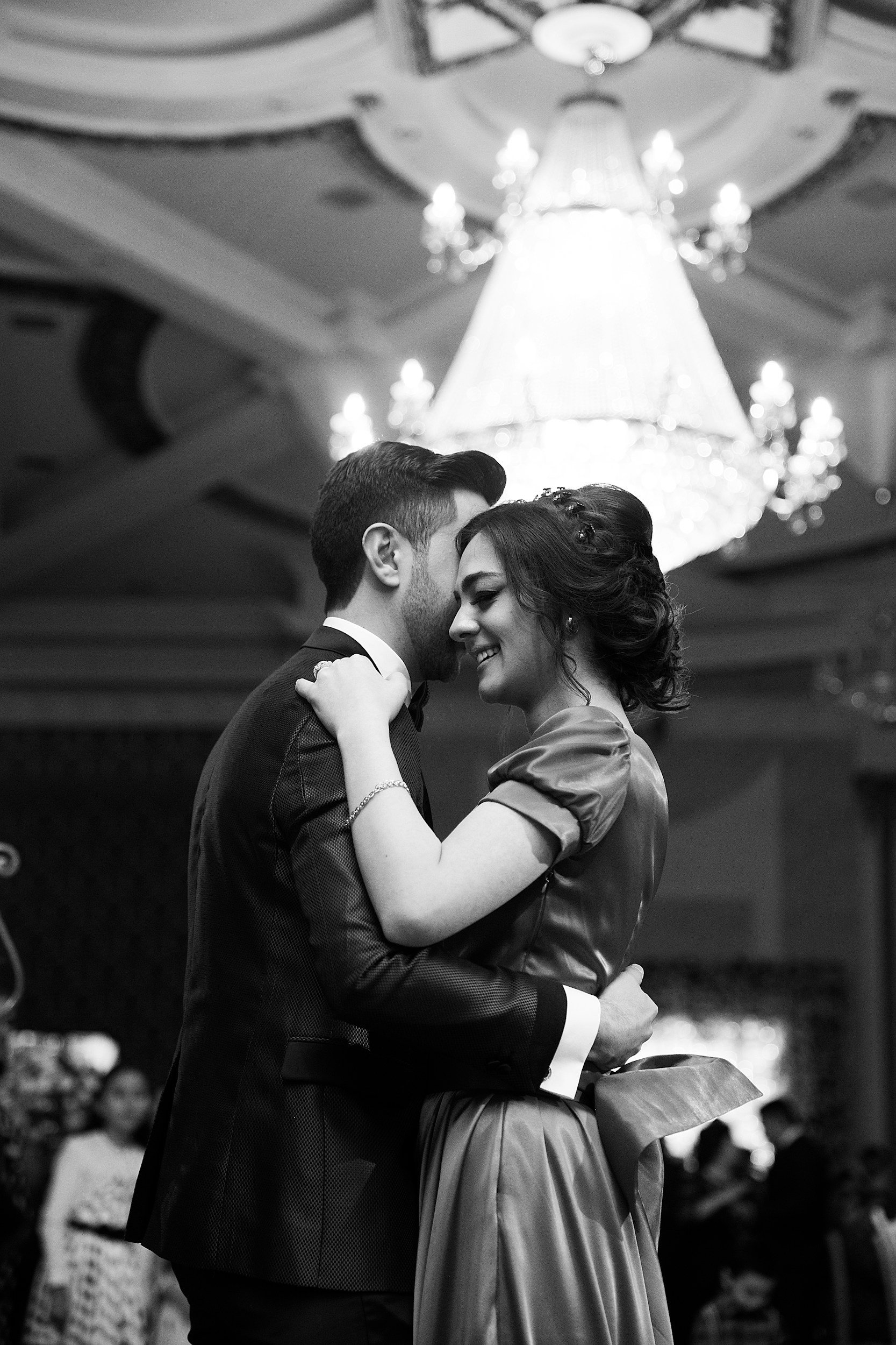 A black and white photo of a smiling couple dancing closely, with a chandelier hanging above them.