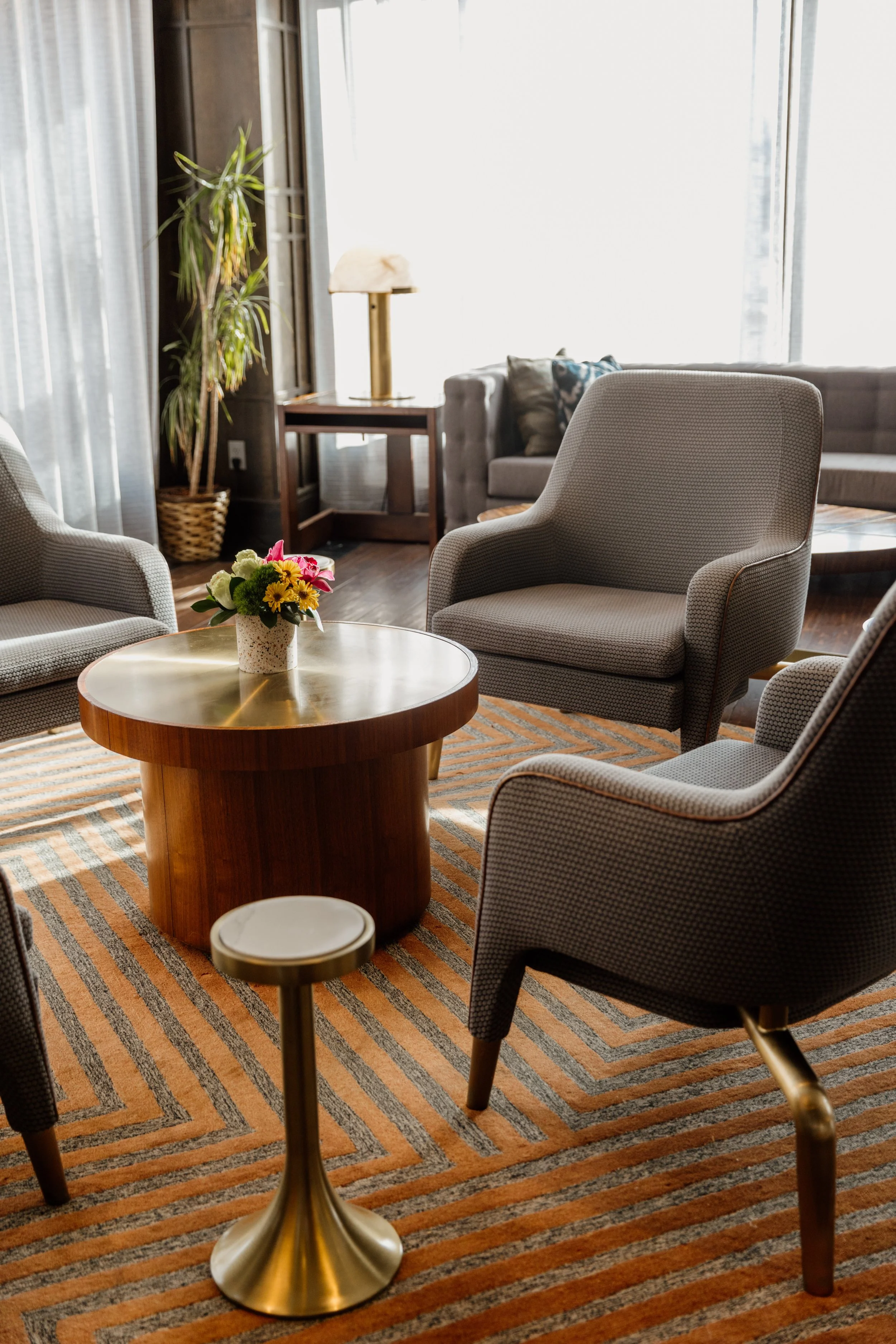 Cozy living room with gray upholstered armchairs, a wooden coffee table with a flower vase, a striped rug, a tall plant, a side table with a lamp, and a sofa with cushions, illuminated by natural light from large windows.
