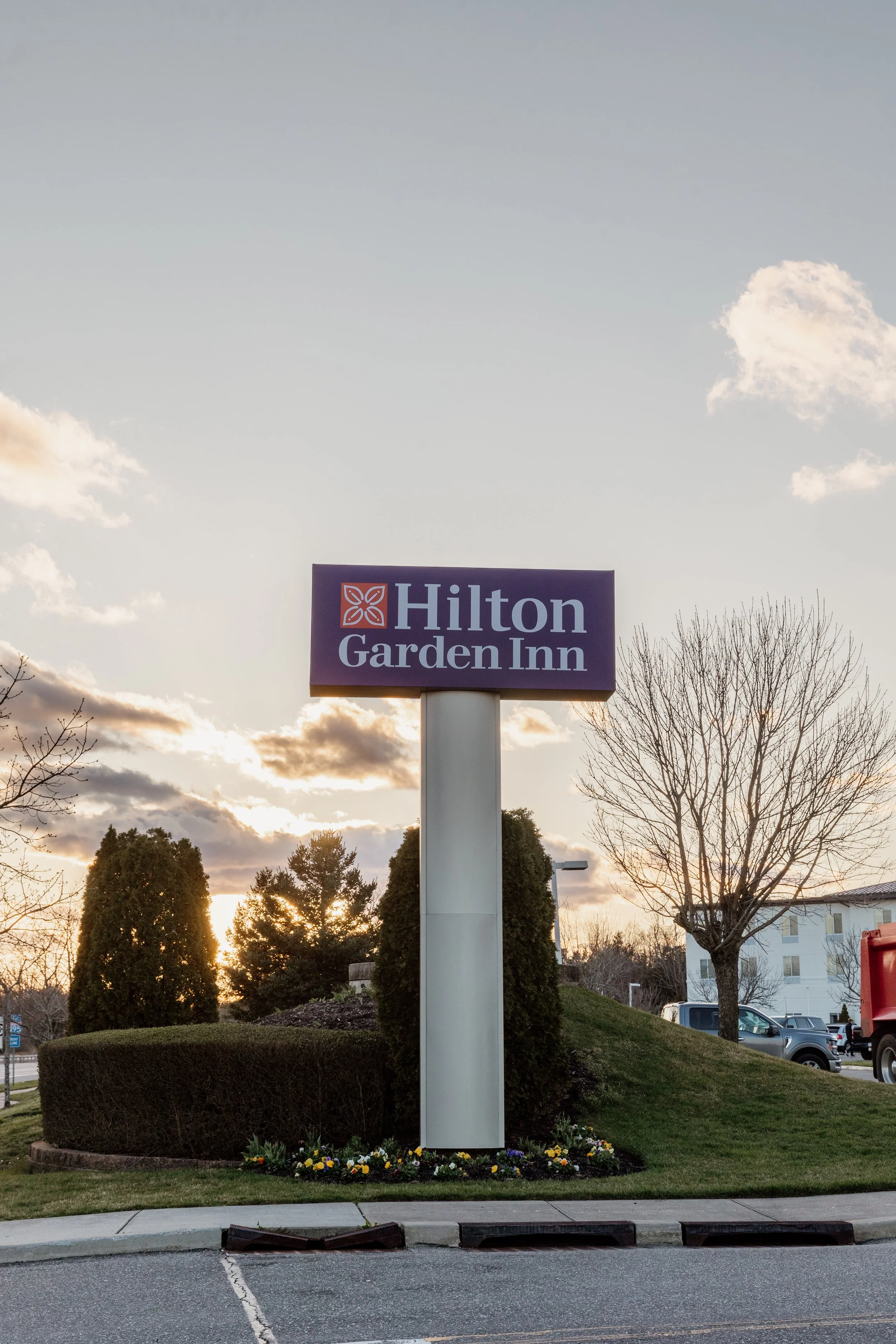 A sign for Hilton Garden Inn hotel with a sunset sky in the background, surrounded by trees and landscaping.