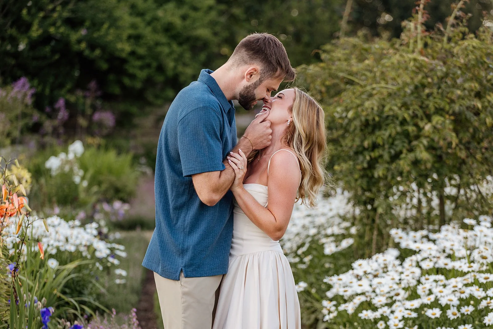 A newly engaged couple caught in a candid, joyful embrace at Harkness Memorial State Park, faces close and smiling, surrounded by garden blooms.
