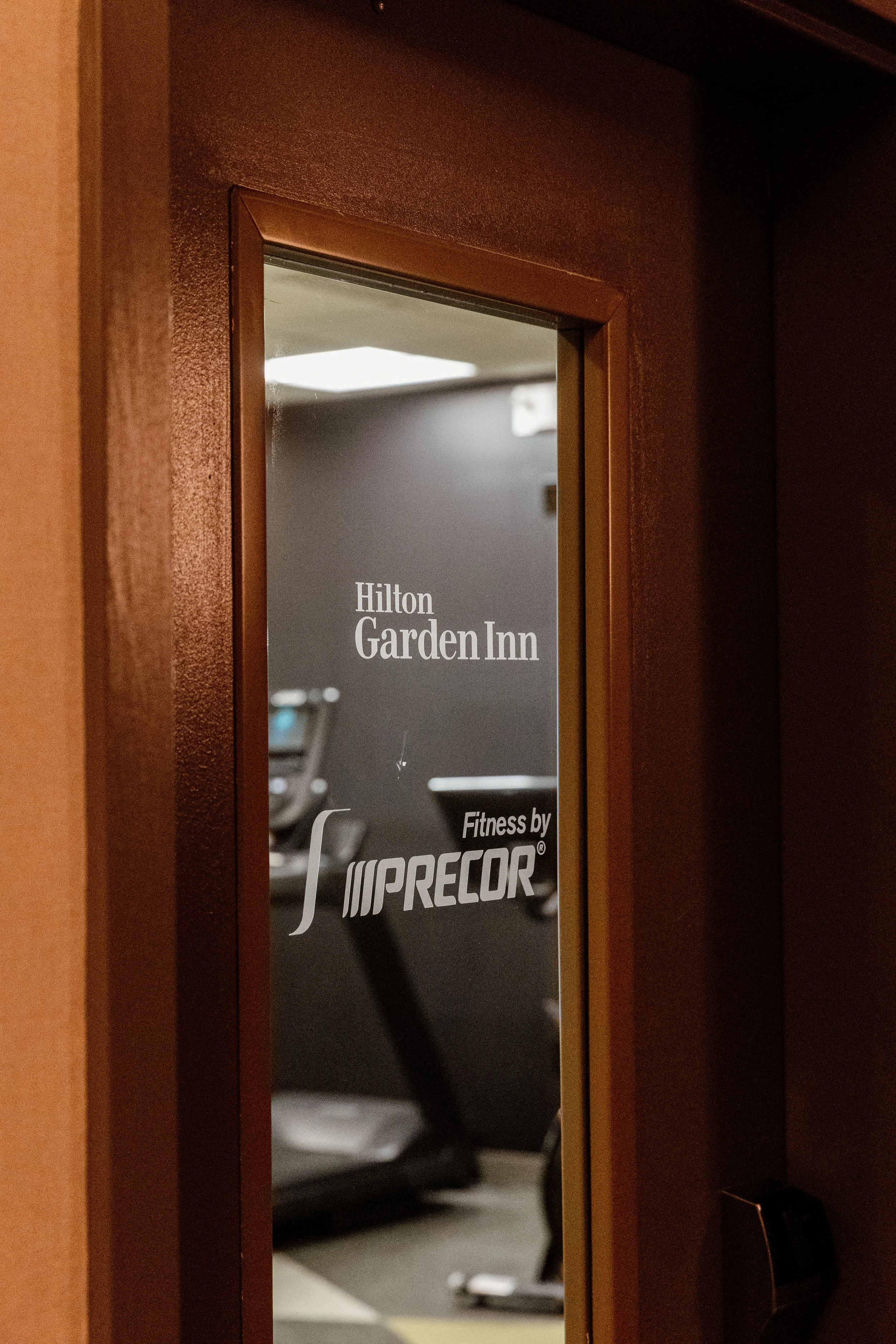 A mirror on a door with the words 'Hilton Garden Inn' and 'Fitness by Precor' reflecting a fitness room with exercise equipment.