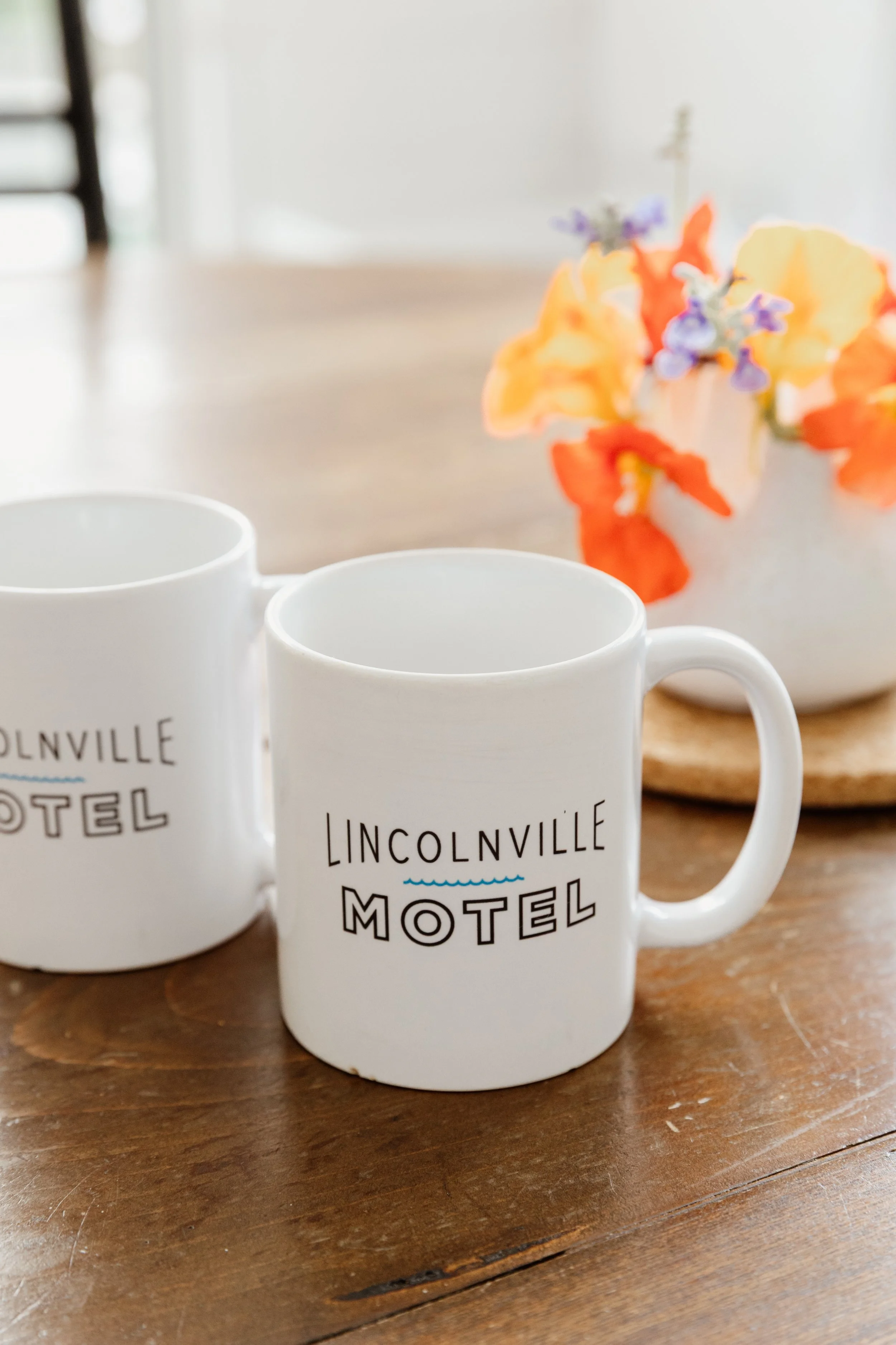 Two white ceramic mugs with 'Lincolnville Motel' printed on them sitting on a wooden table, with a flower arrangement in a white vase in the background.