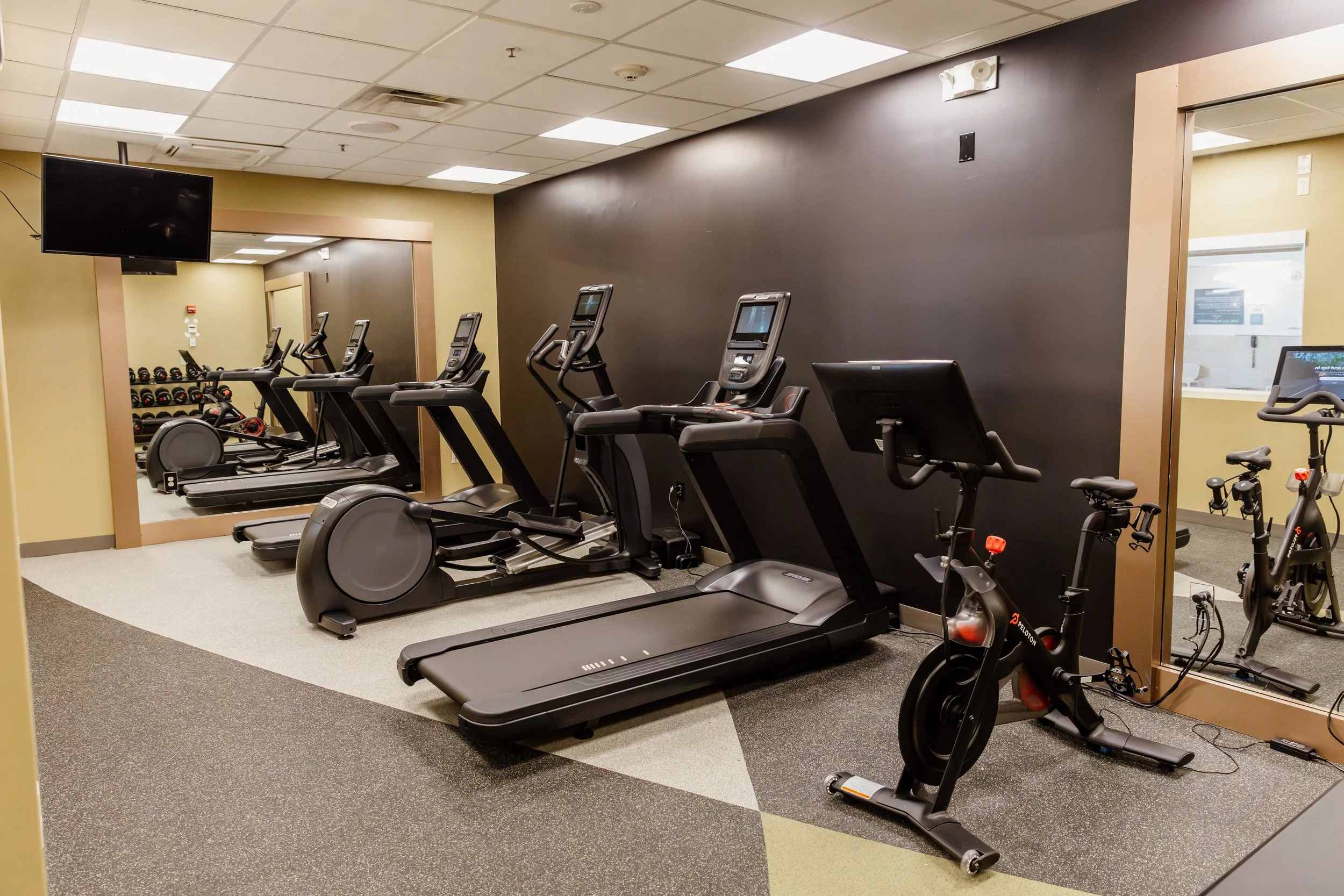 A small gym room with exercise equipment including treadmills, elliptical machines, and stationary bikes, with a wall mirror and a TV mounted on the wall.