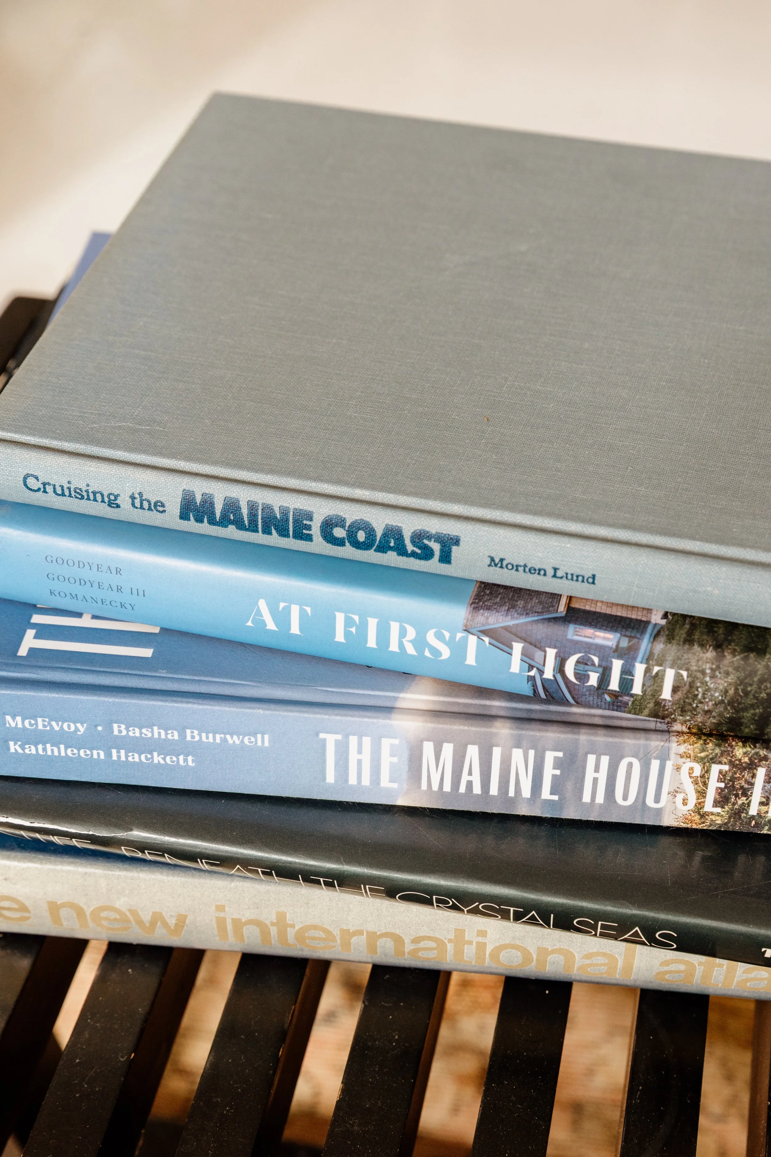 Stacked books about Maine, including 'Cruising the Maine Coast,' 'At First Light,' 'The Maine House,' and 'The New International Atlas,' on a wooden table.
