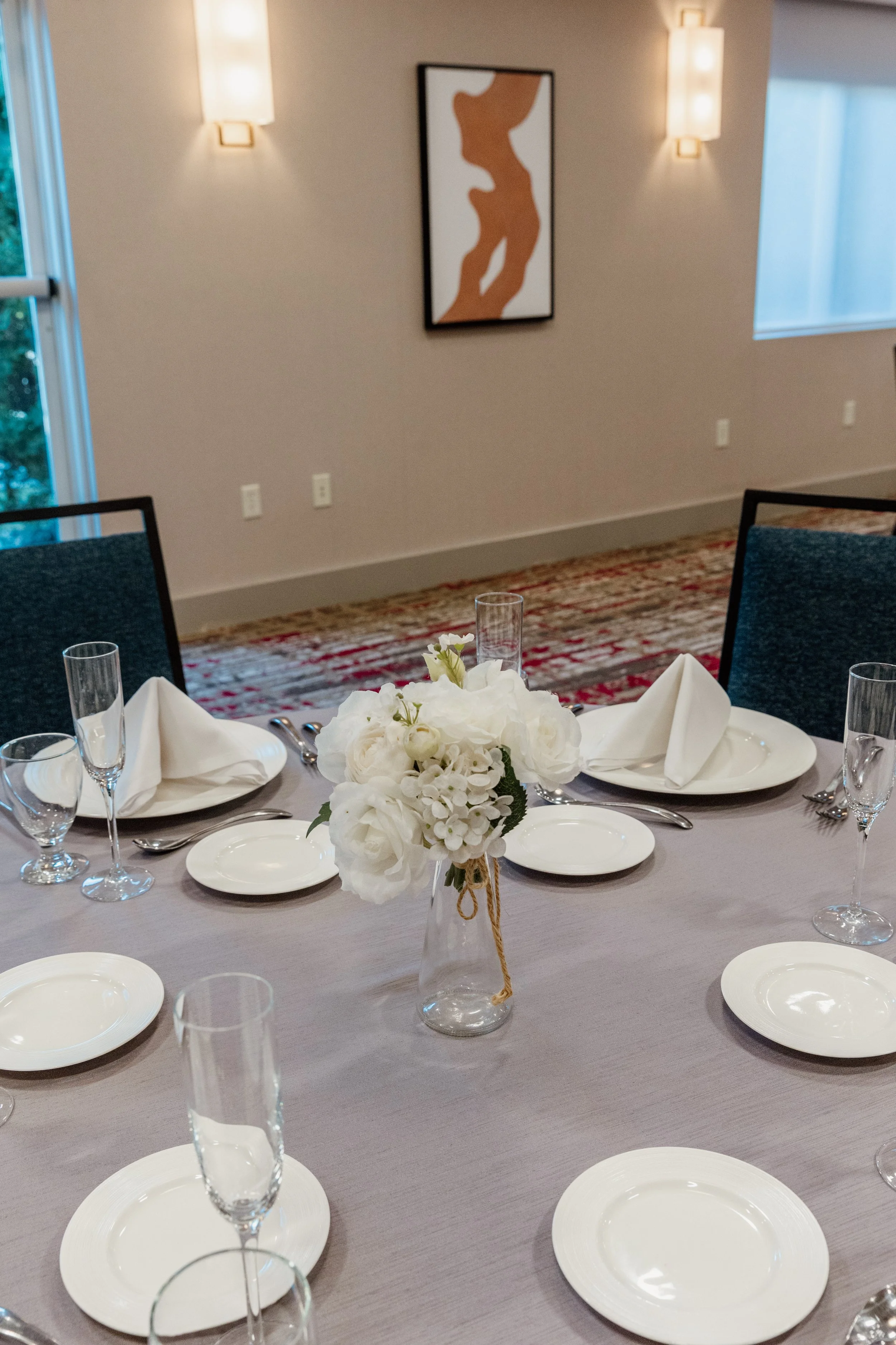 Round dining table set for a formal event with white plates, folded napkins, wine glasses, and a vase with white flowers at the center, in a room with wall art and wall-mounted lights.