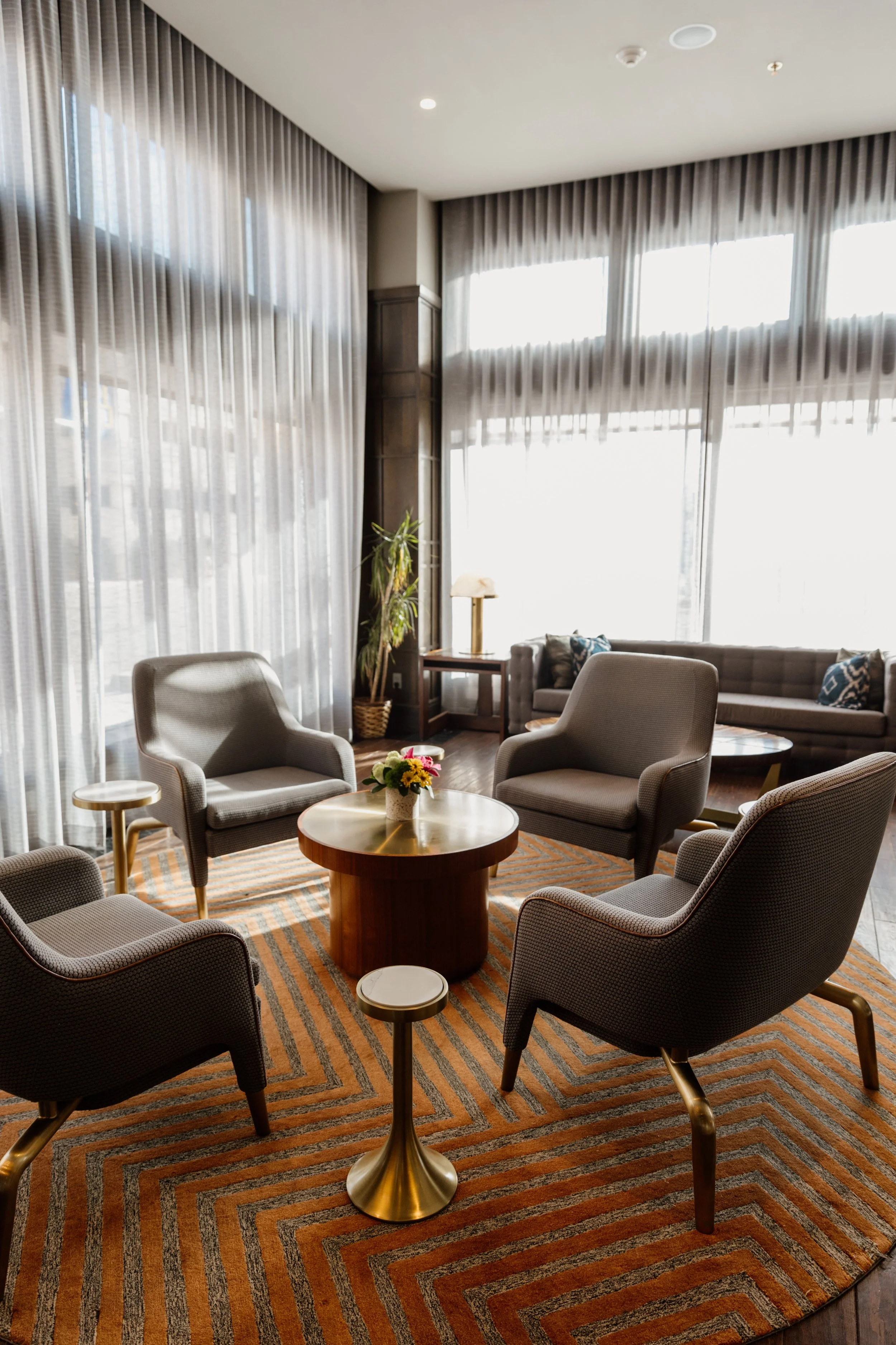 Cozy hotel lobby area with four armchairs around a round coffee table, a flower arrangement on the table, and large windows with sheer curtains letting in natural light.