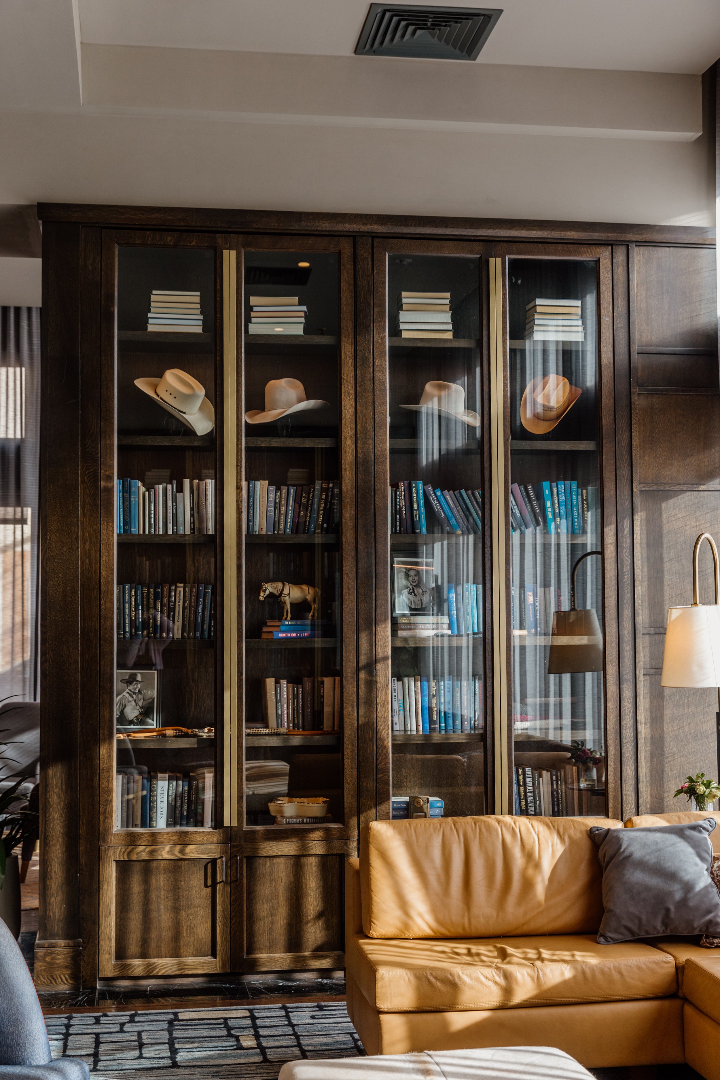 Wooden bookshelf with glass doors filled with books, hats, and framed photographs, located in a cozy living room with a tan leather sofa and modern lighting.