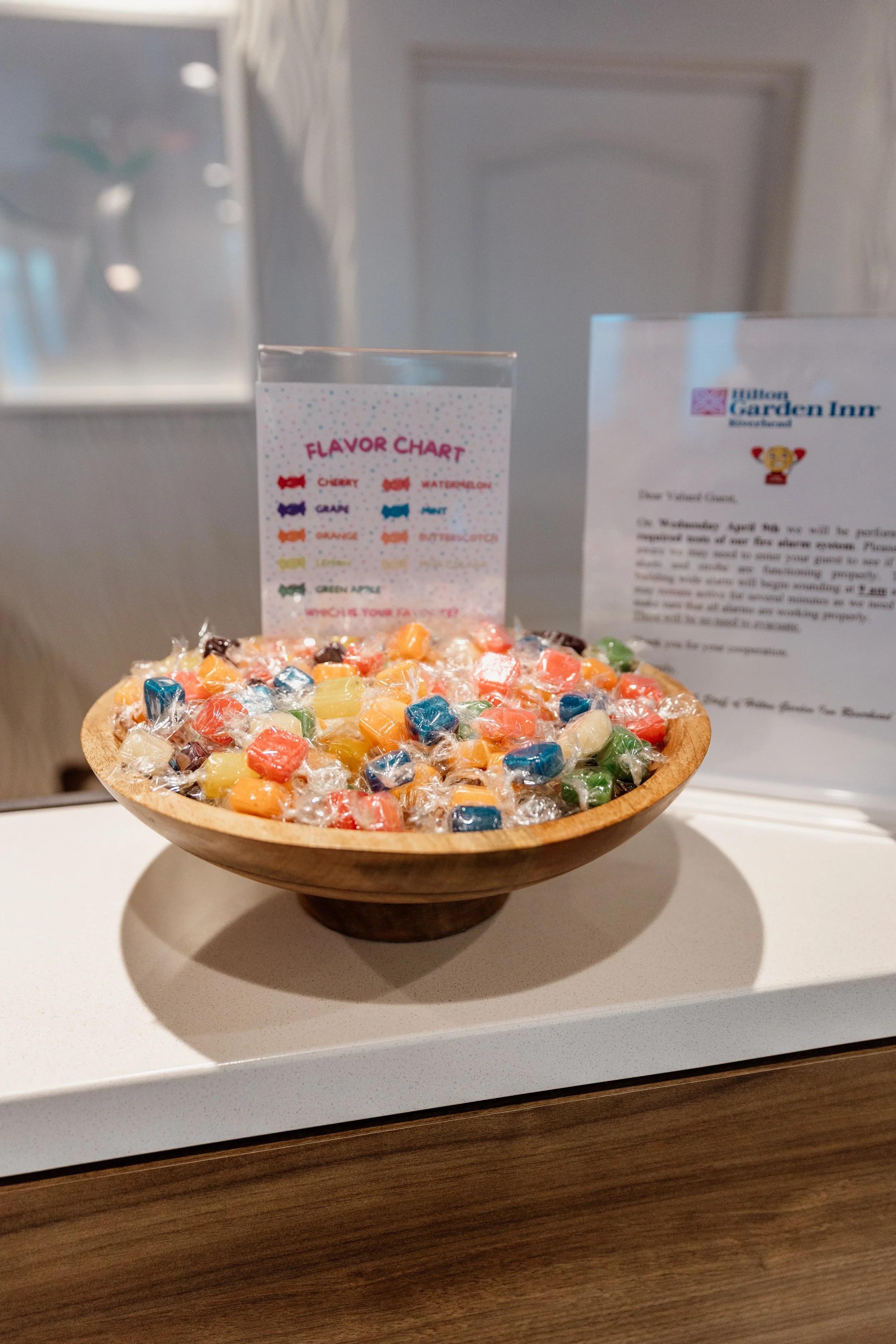 A wooden bowl filled with assorted wrapped candies on a white surface, with a flavor chart and a sign for Hilton Garden Inn in the background.