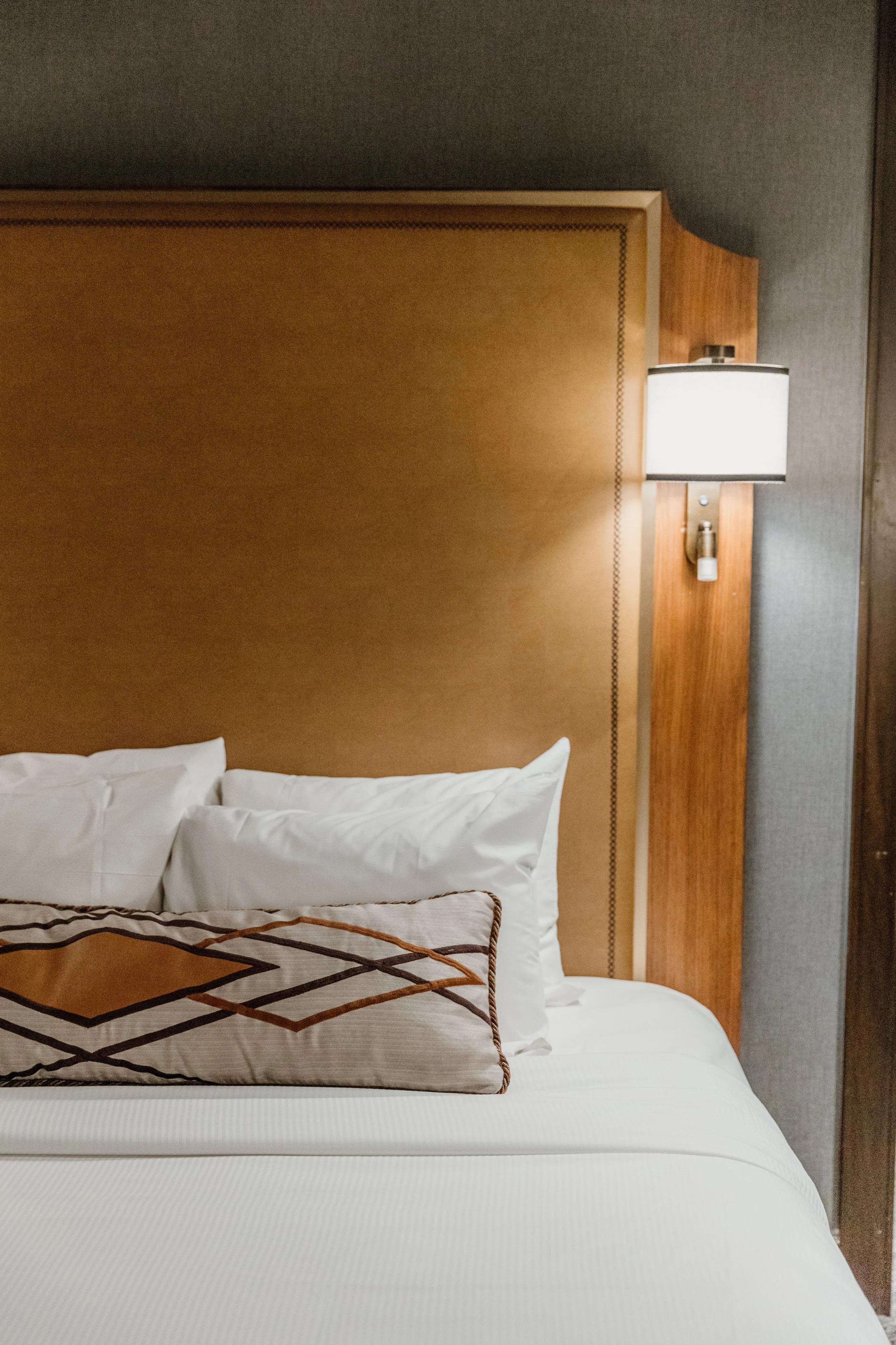 A cozy hotel bed with crisp white pillows and a decorative accent pillow against a warm wooden headboard, captured during a content business shoot at the Graduate by Hilton in Fayetteville, Arkansas.