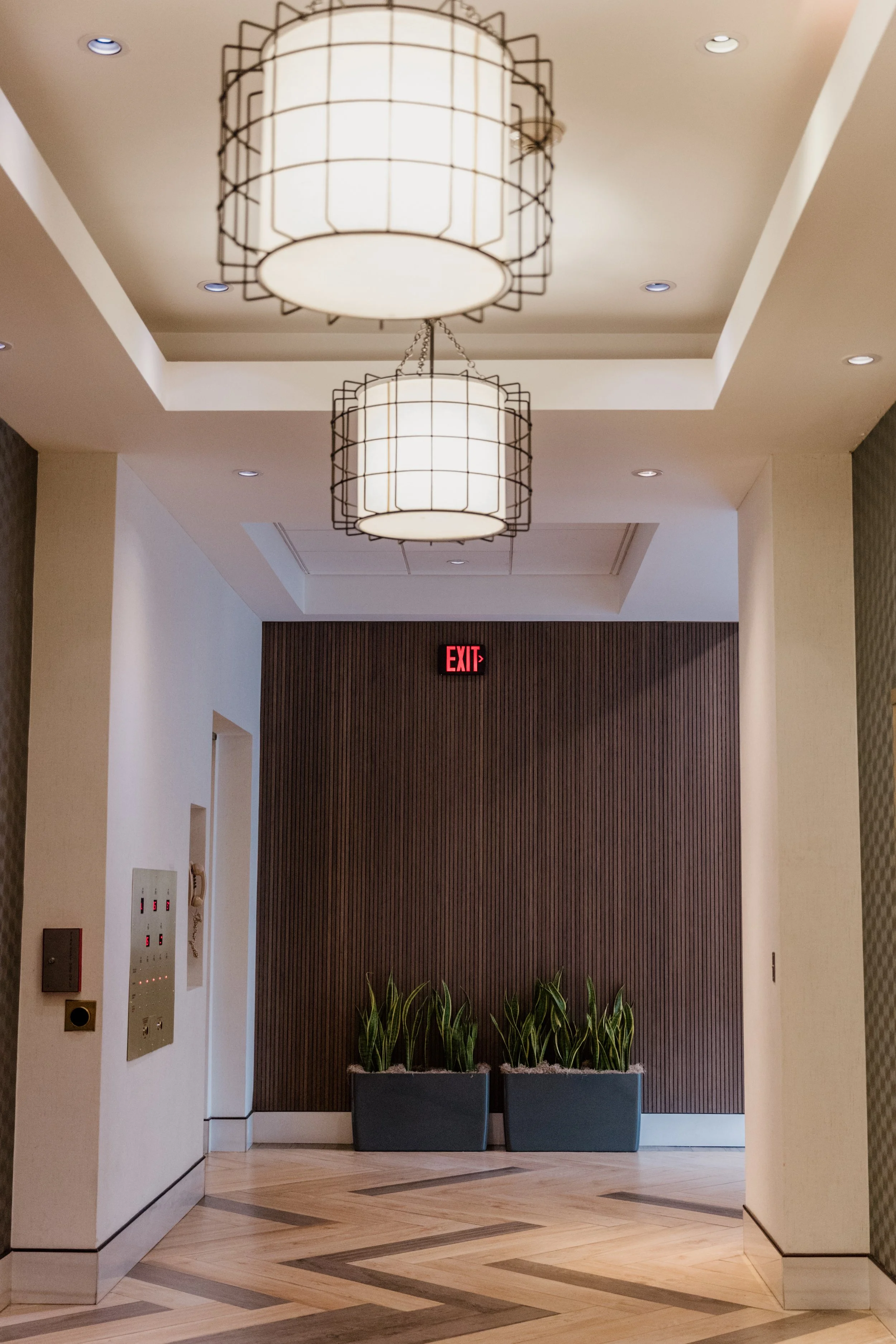 Lobby with a dark wood accent wall, two large hanging light fixtures, an illuminated red exit sign, and two planters with green plants on a light-colored wood floor with a geometric pattern.