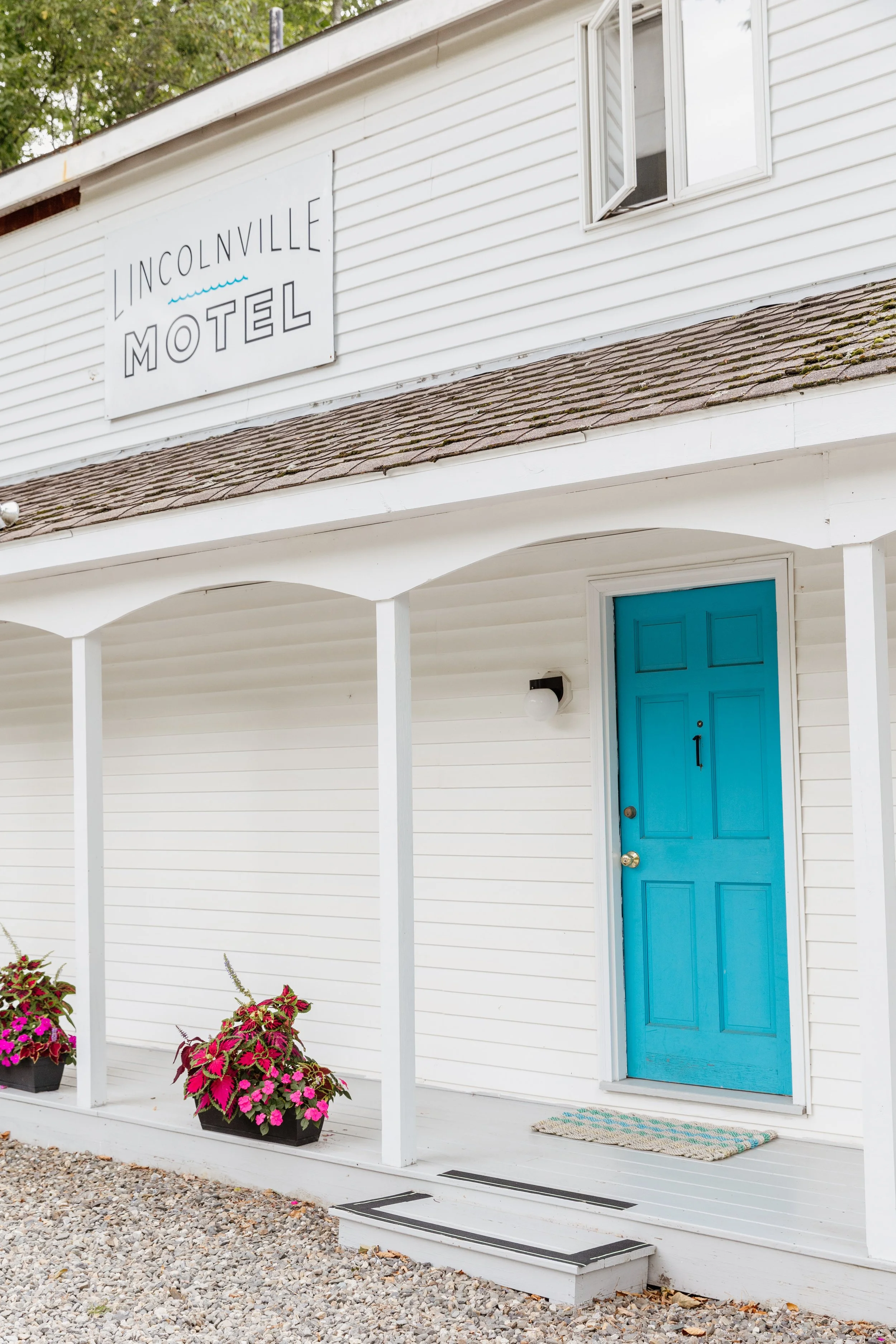 Exterior of Lincolnville Motel with a white building, bright blue front door, potted flowers, and a sign reading 'Lincolnville Motel'.