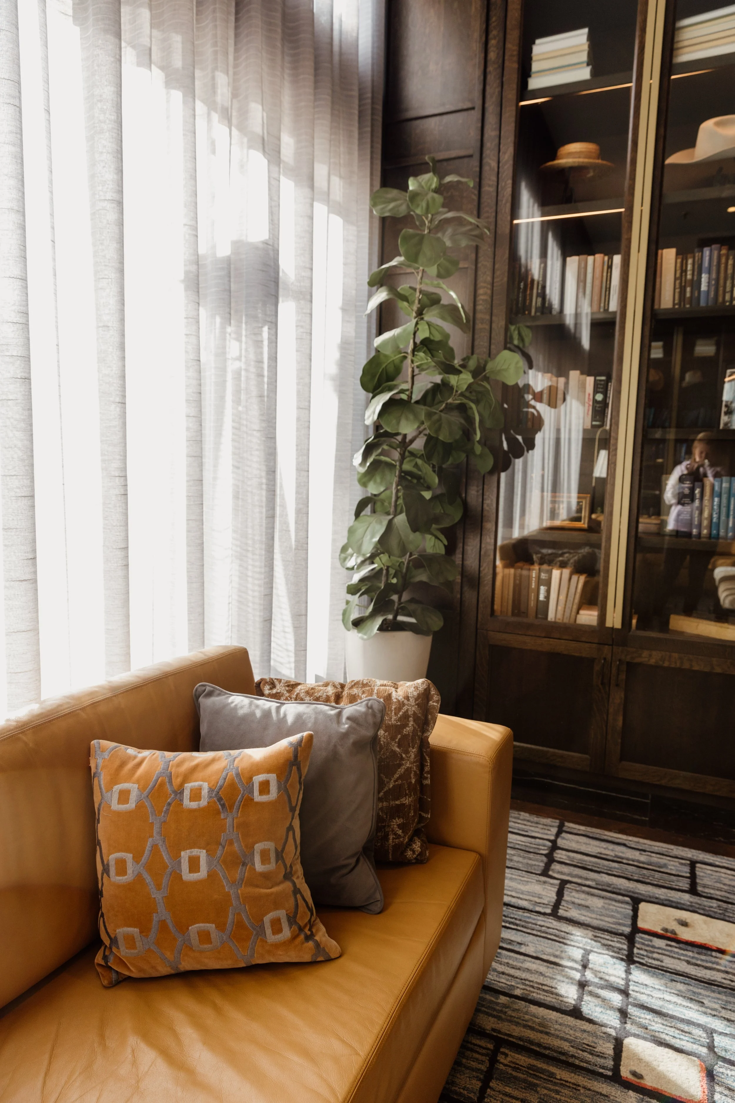 A bright and cozy lounge corner with a beige leather sofa, layered decorative pillows, a tall leafy plant, and a glass-front bookshelf, captured during a content business shoot at the Graduate by Hilton in Fayetteville, Arkansas.