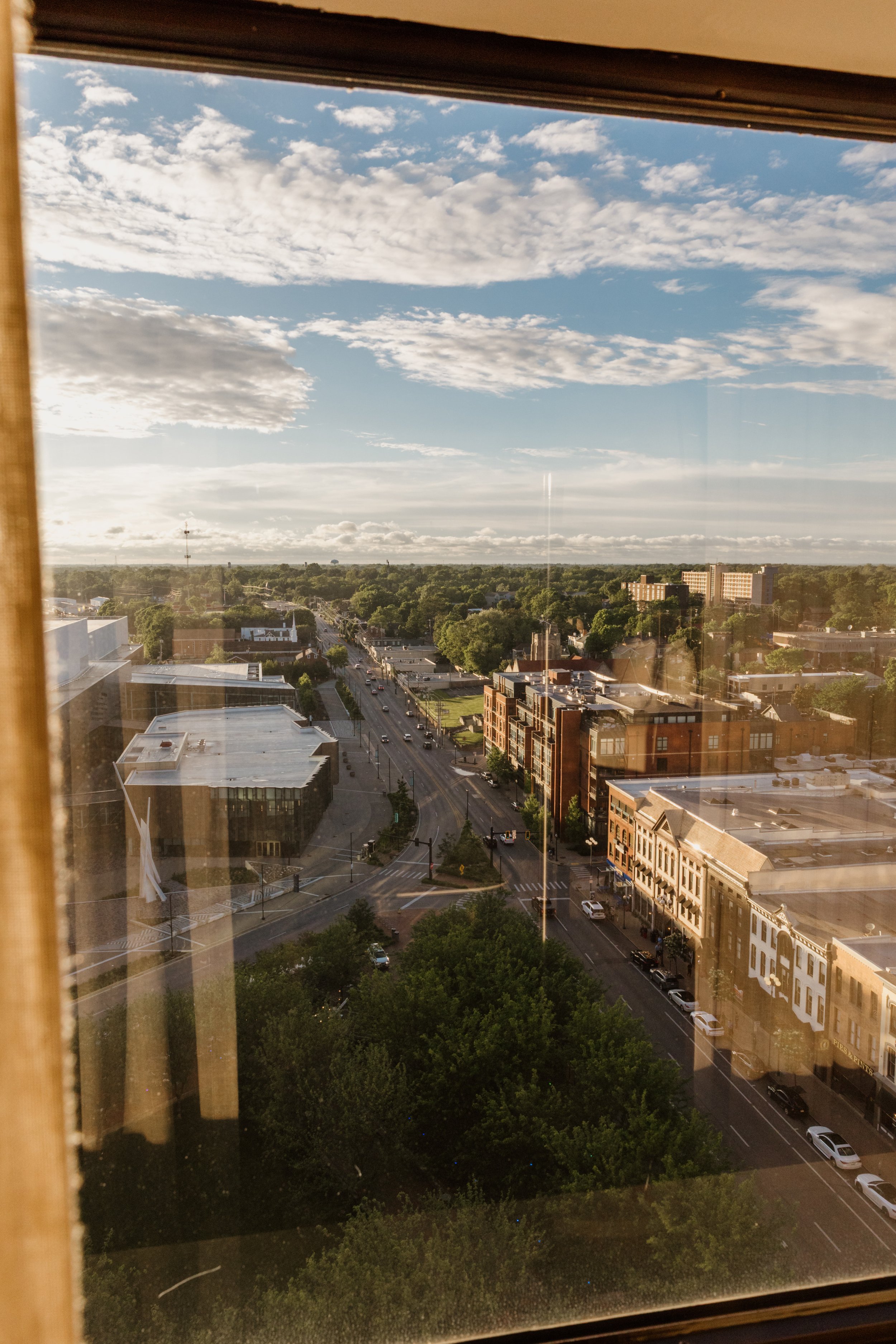 View of a downtown city street from a high window, with buildings, trees, and a partly cloudy sky.